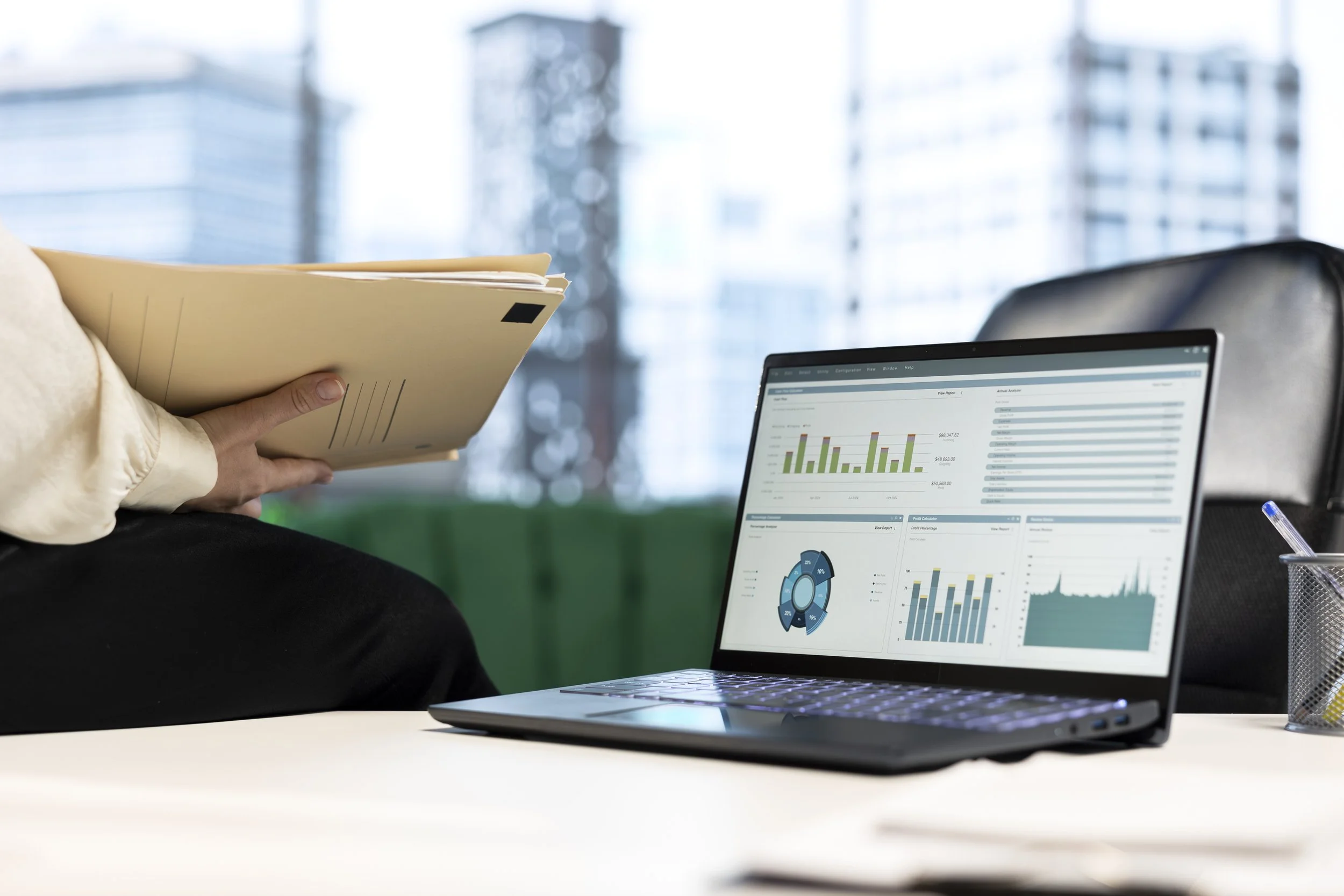 Person holding a folder while working with a laptop displaying graphs and charts, with a cityscape through a large window in the background.