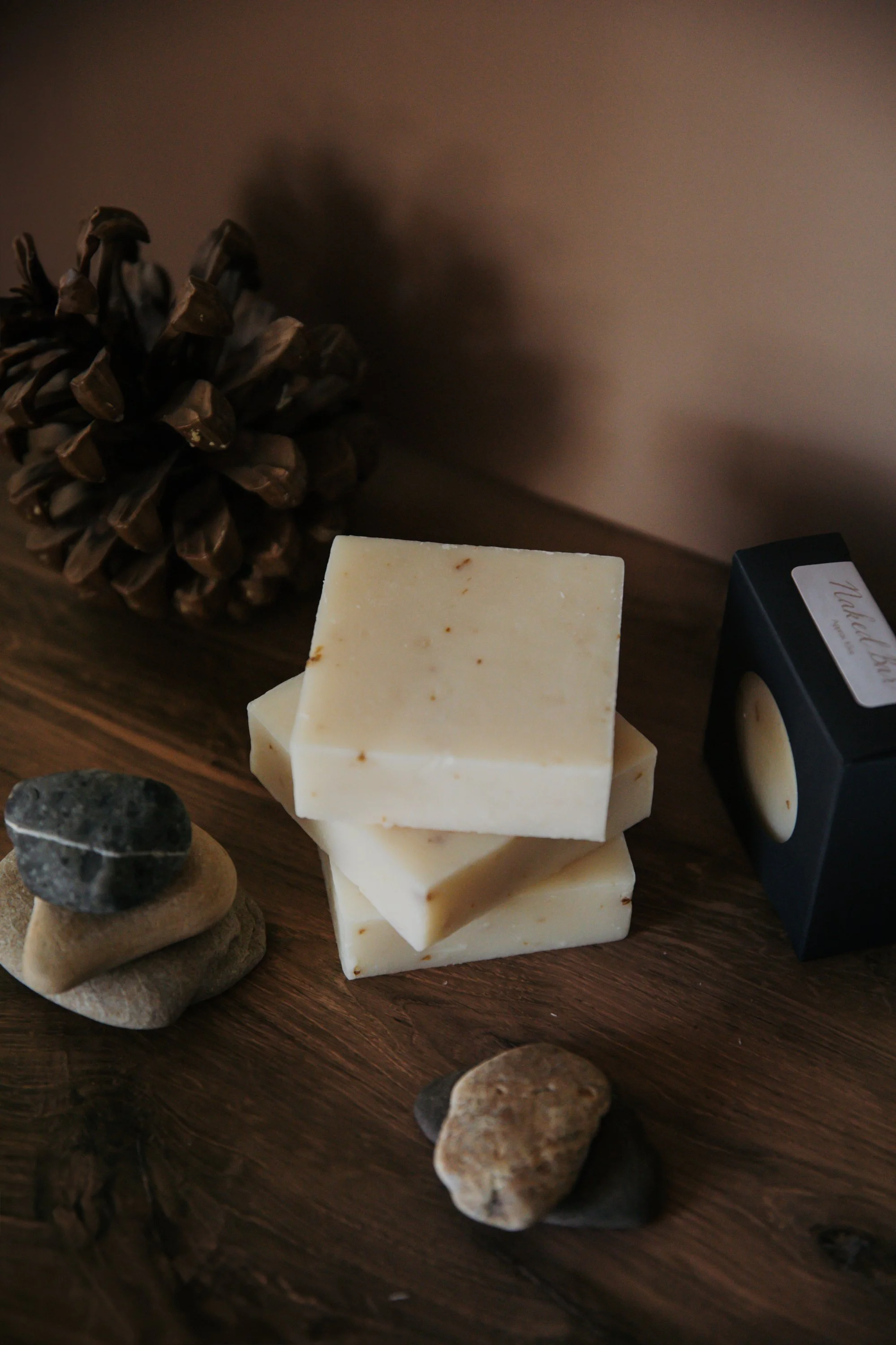A pinecone, a stack of white soap bars, stacked stones, and a small black box with a white label on a wooden surface.