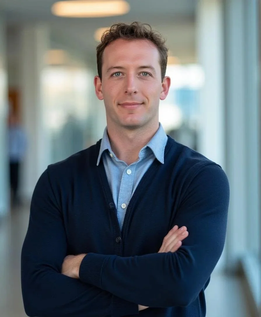A man stands with arms crossed in an indoor setting with large windows and blurred background.