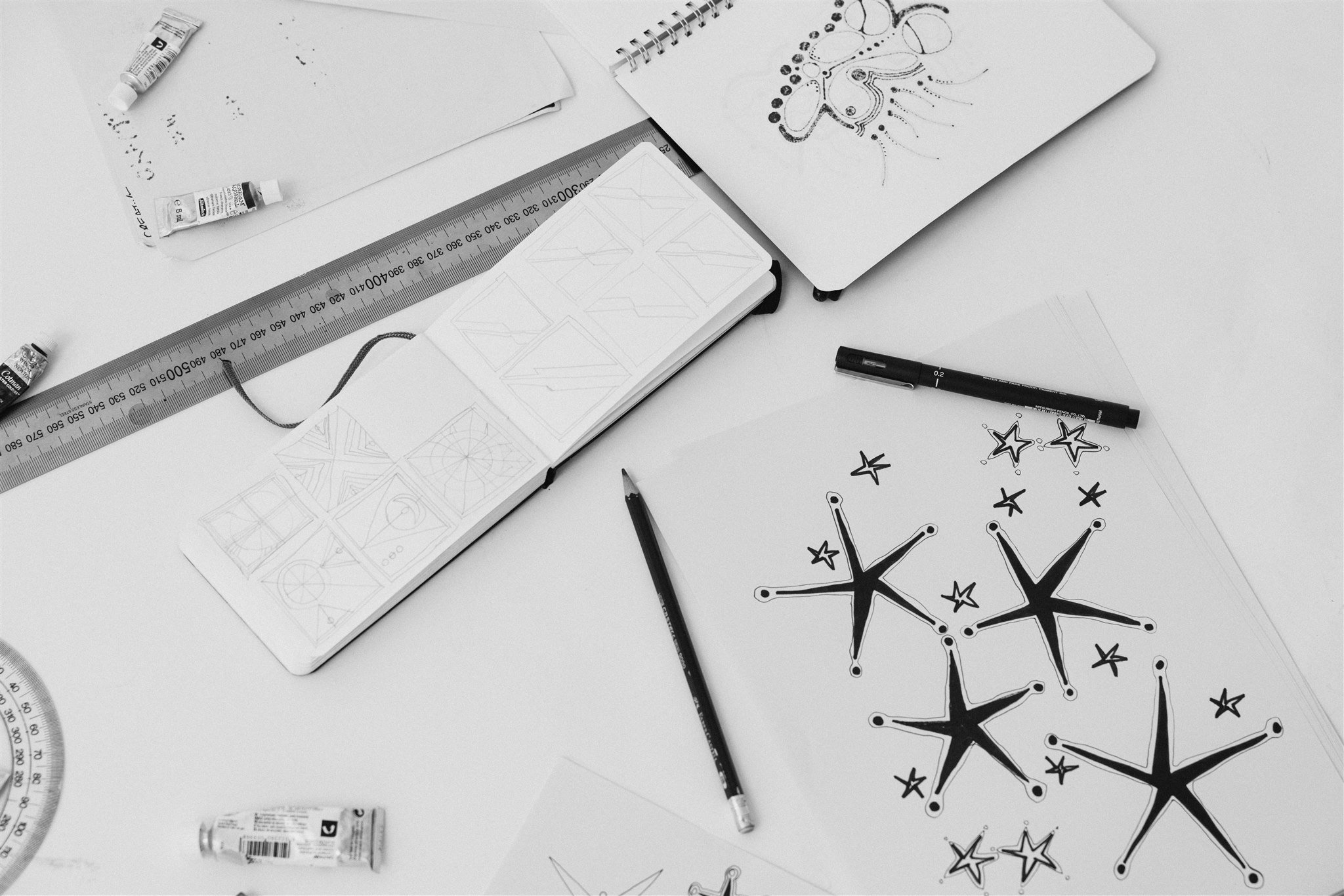 Black and white photo of art supplies including a ruler, pens, markers, and sketches of stars and abstract designs on paper spread on a white table.