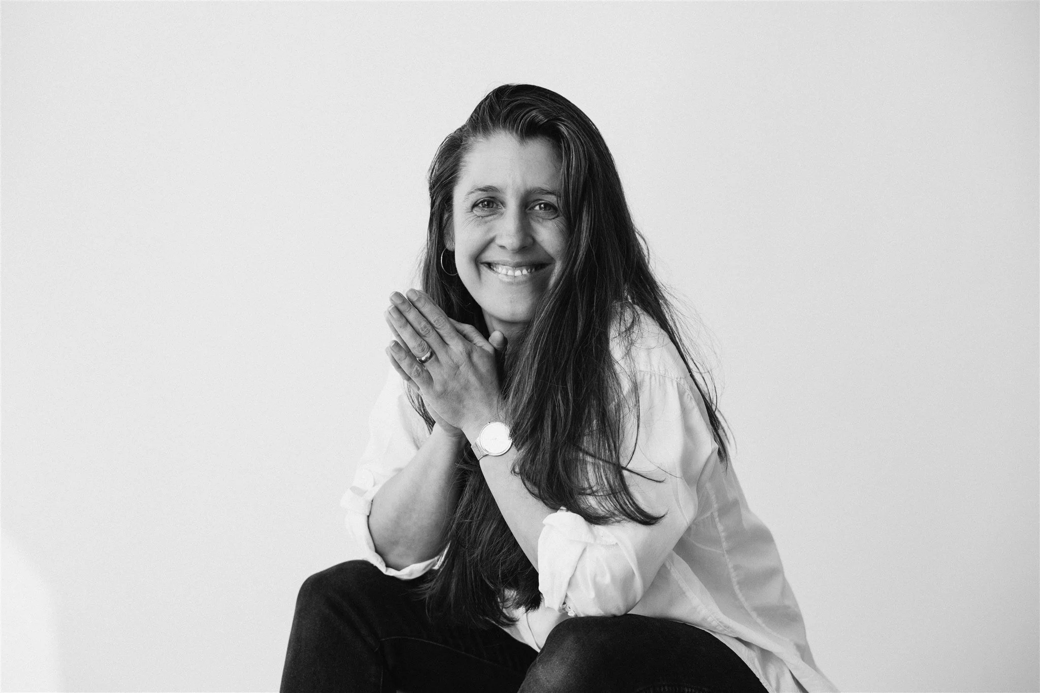 Black and white portrait of Kate smiling, wearing hoop earrings, a watch, and casual clothing, sitting against a plain white background.