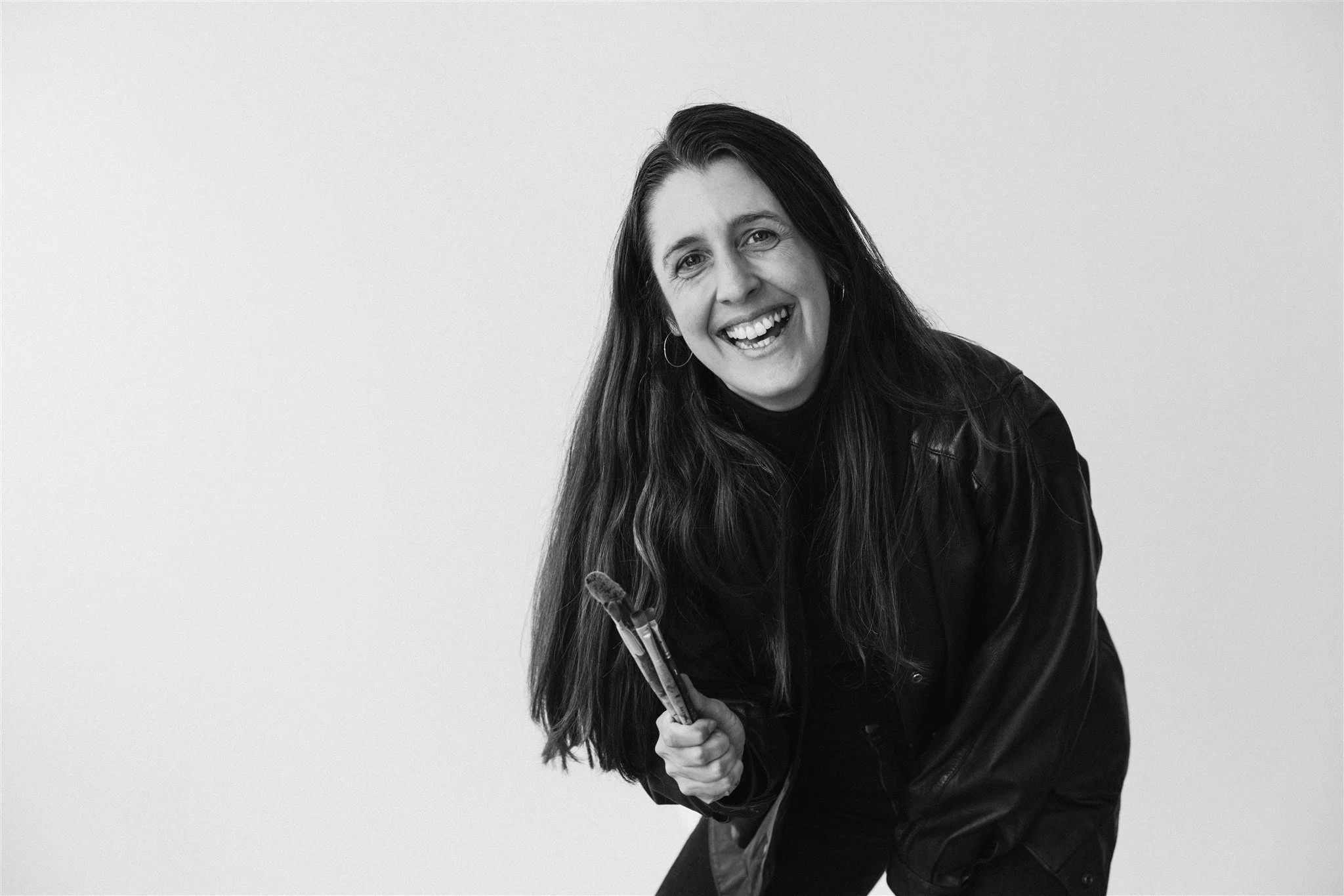 Black and white photo of Kate smiling and holding a paintbrush in her hand, wearing a black leather jacket, against a plain light background.