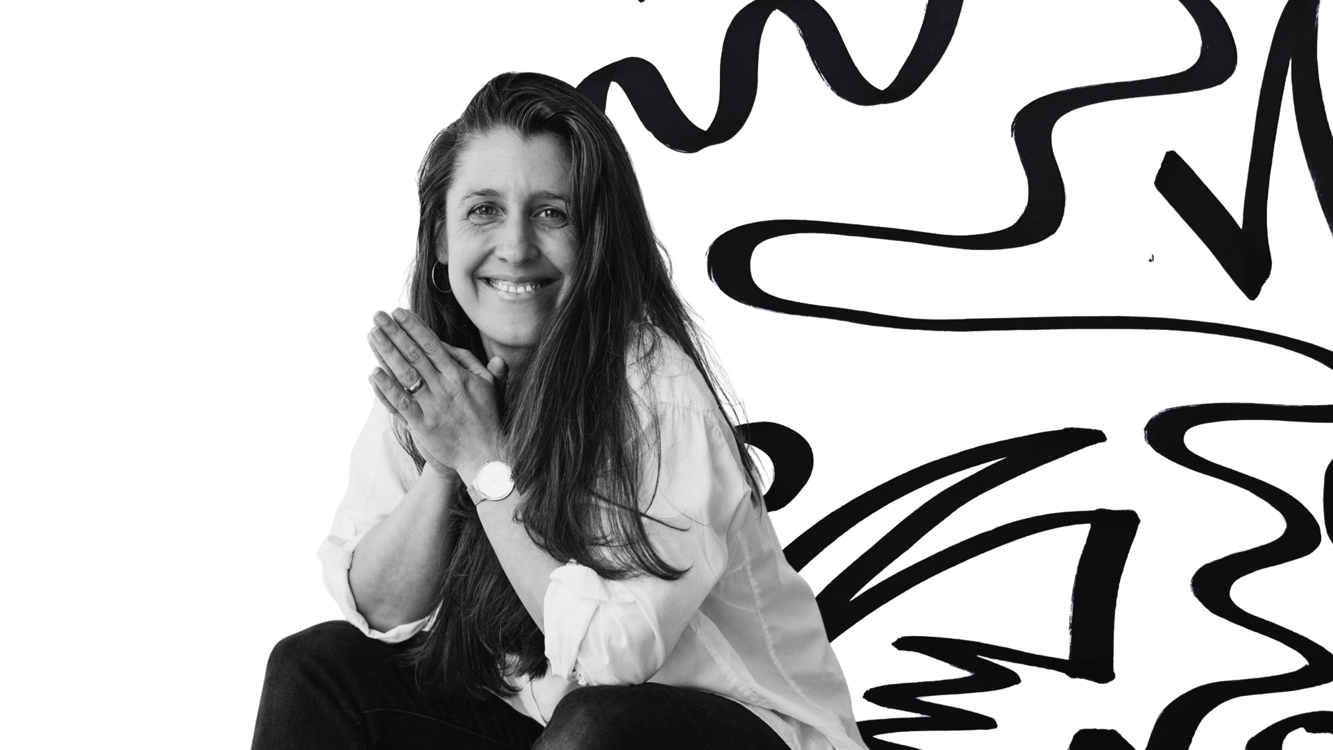 Black and white photo of a woman with long hair smiling, sitting with her hands clasped near her face, wearing a watch and earrings, with abstract black lines and shapes in the background.