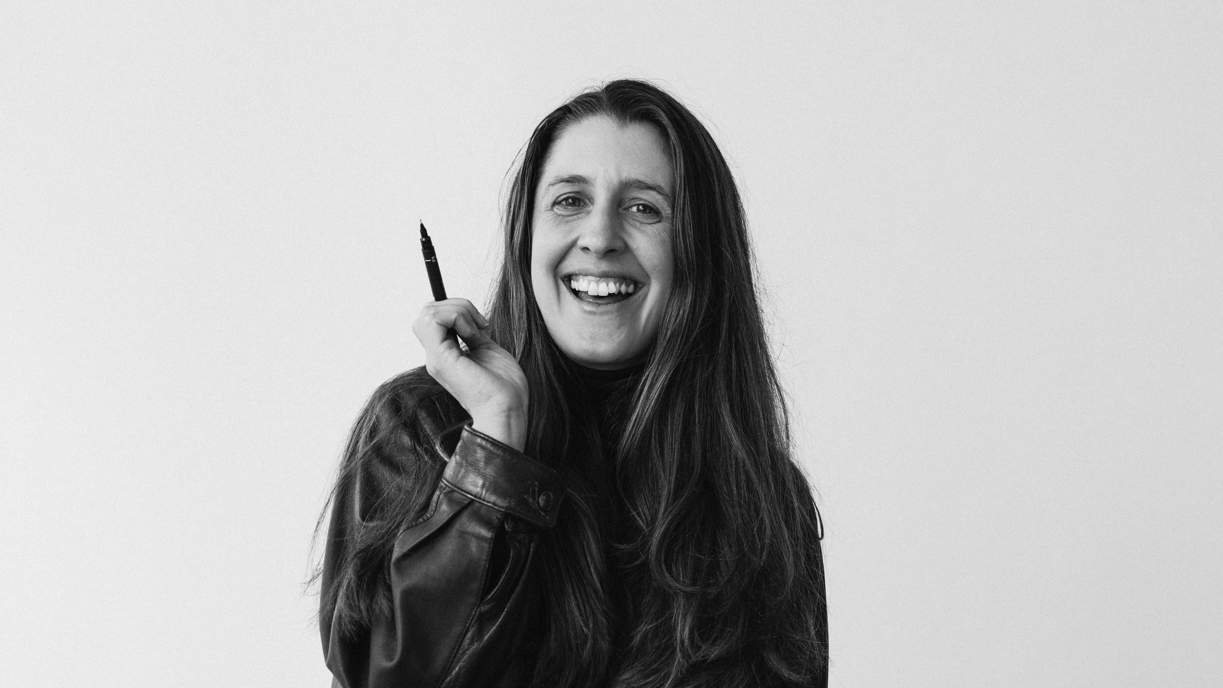 Black and white photo of Kate smiling, wearing a leather jacket, holding a pen, standing against a plain background.