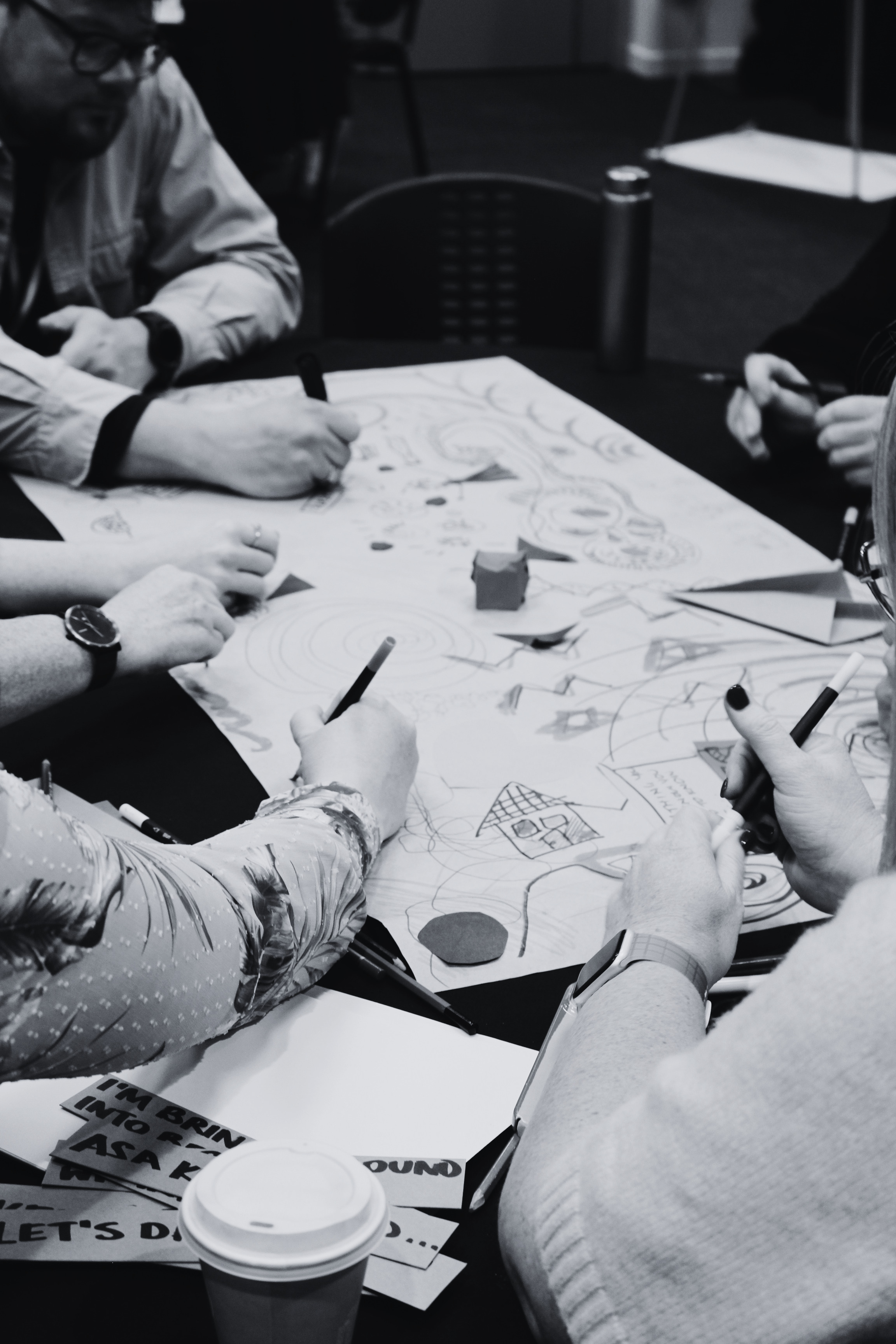 A group of people are working together on a large sheet of paper with drawings and notes. Several hands are visible holding pens, and there is a coffee cup on the table. The photo is in black and white.