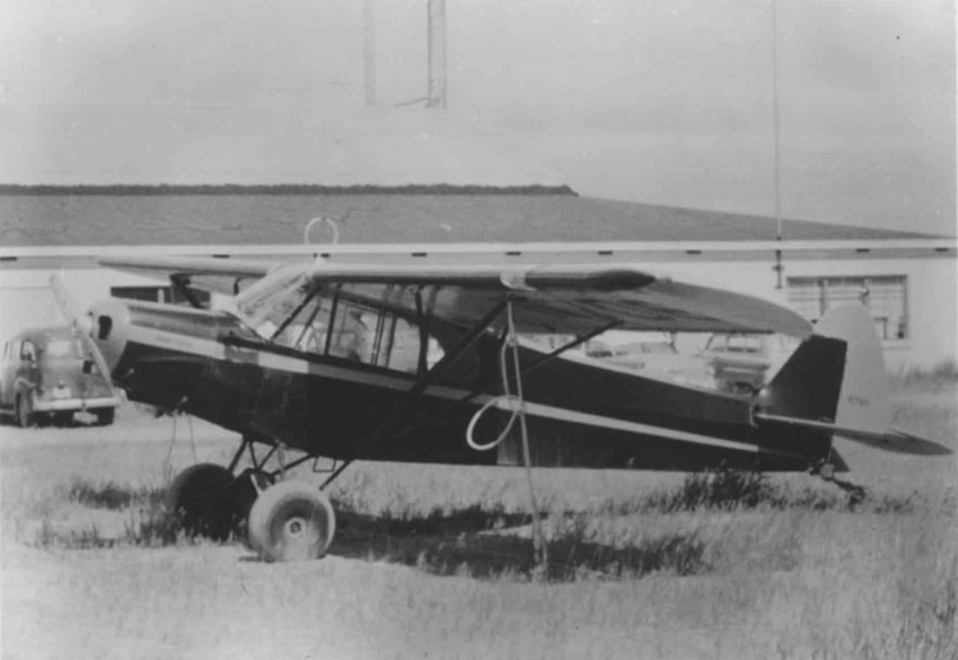 Black and white photo of a small airplane on grass near a hangar, with an older car parked nearby.