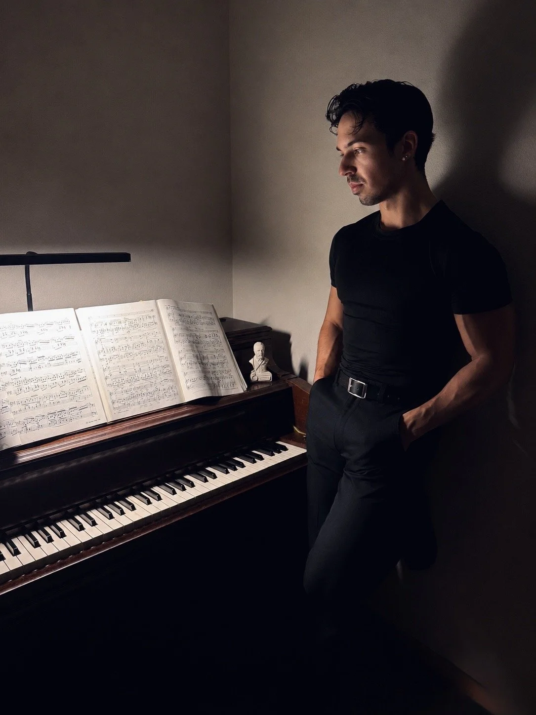 A young man with dark hair, dressed in black, leaning against a wall next to an open piano with sheet music, a small bust, and a music stand on top.