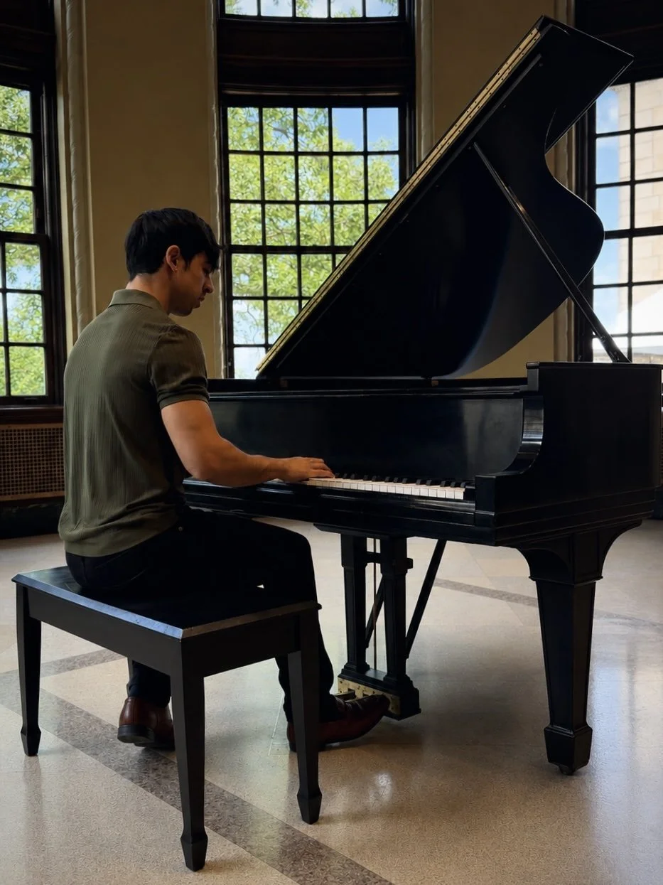 A man playing a grand piano in a room with large windows and natural light.