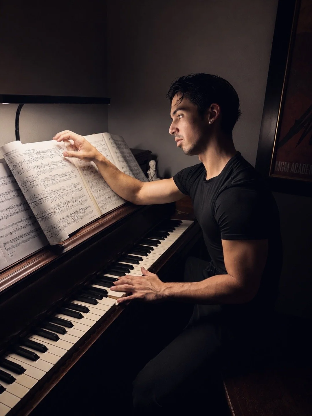A man playing a piano in a dimly lit room, with sheet music open on the piano and a poster on the wall.