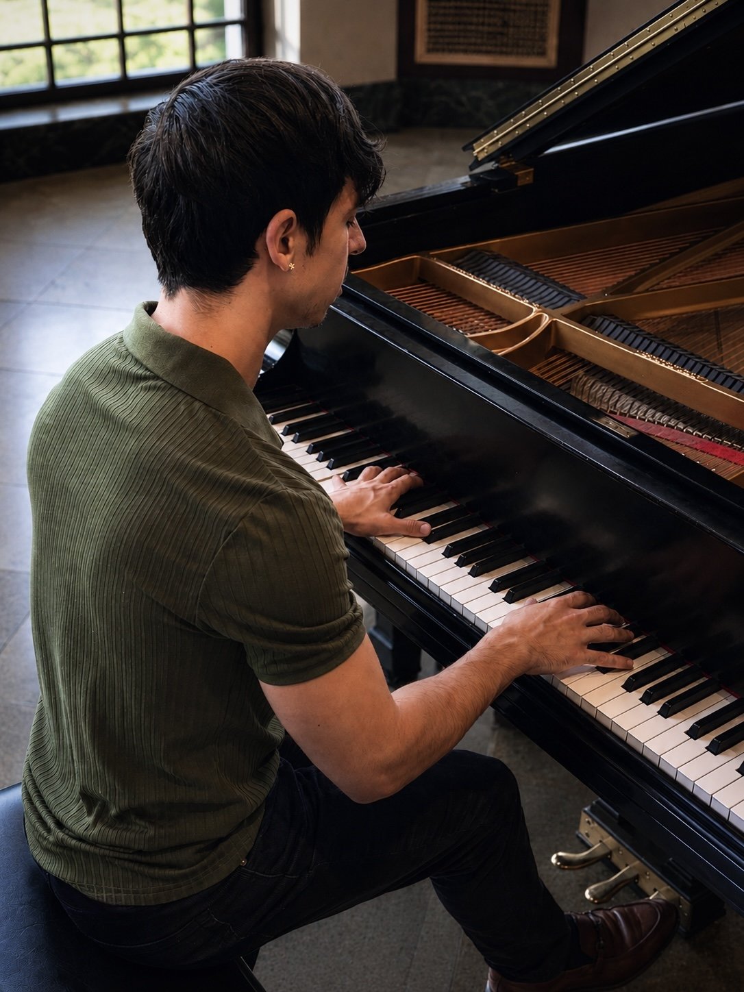 A man playing a black grand piano in a room with large windows.