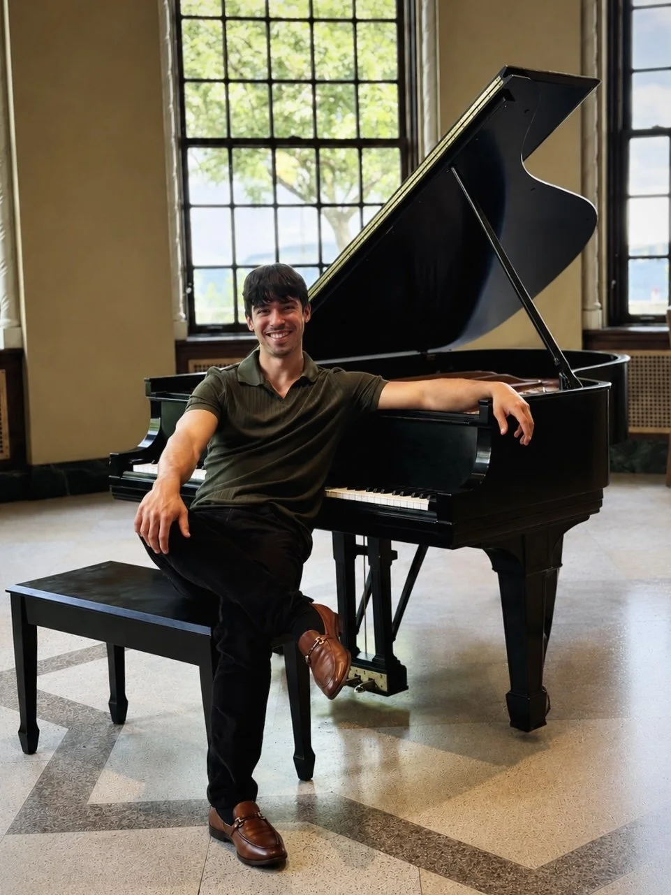 Man sitting at a black grand piano in a room with large windows and a view of trees outside, smiling at the camera.