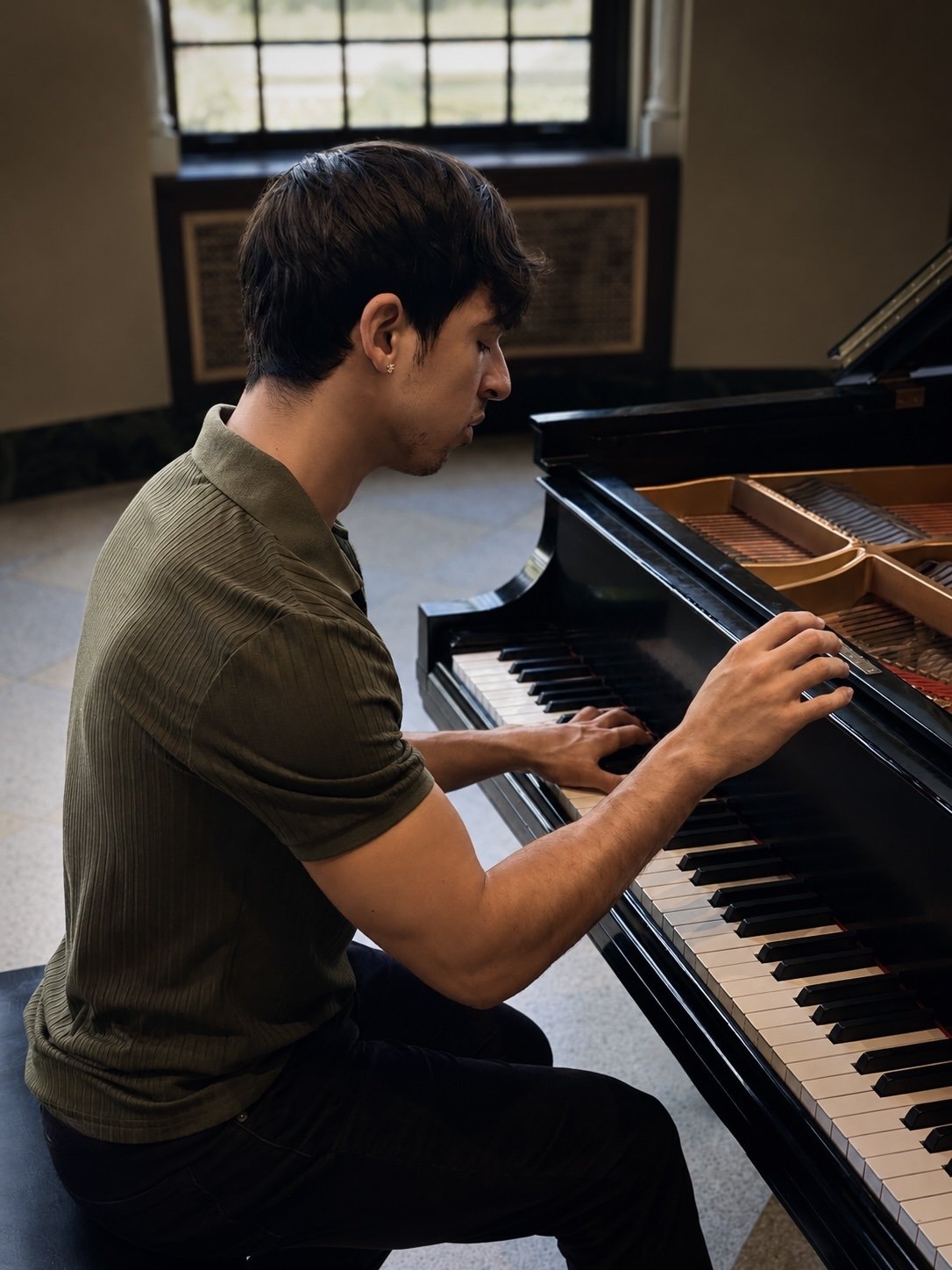 A young man playing a grand piano indoors near a window with a grid pattern.