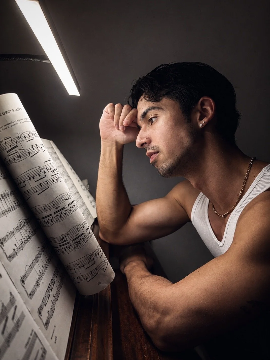 Young man resting his head on his hand, sitting at a piano with sheet music, under a bright overhead light, wearing a white tank top and necklace.