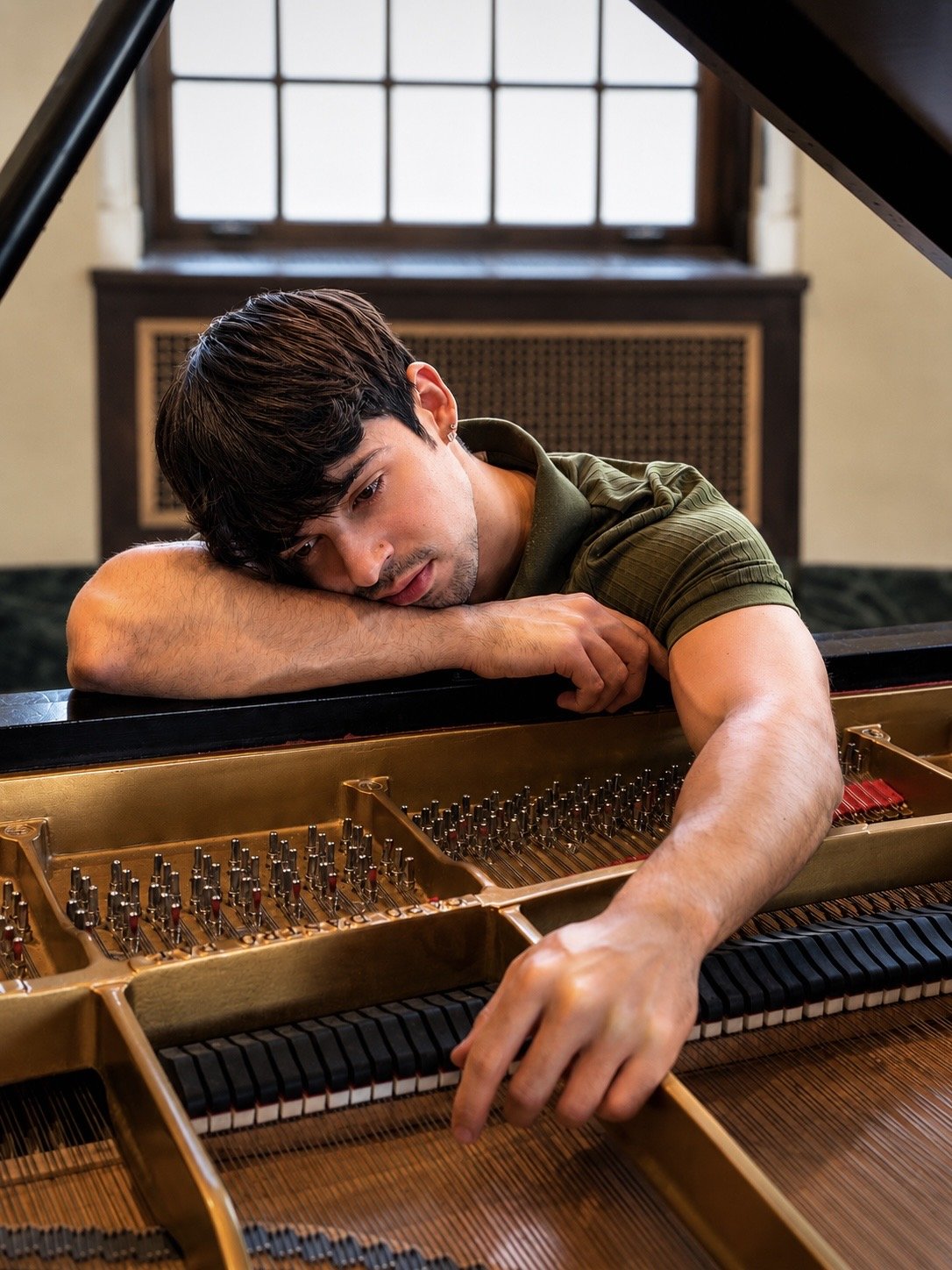 A young man with dark hair resting his head on a grand piano with open lid, in a room with large window and dark wood paneling.
