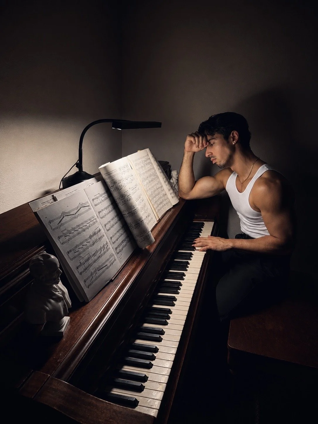 A young man with dark hair, wearing a white tank top, sits at a piano with sheet music. He appears to be deep in thought, resting his head on his left hand with his elbow on the piano. The scene is dimly lit, with a lamp illuminating the sheet music.