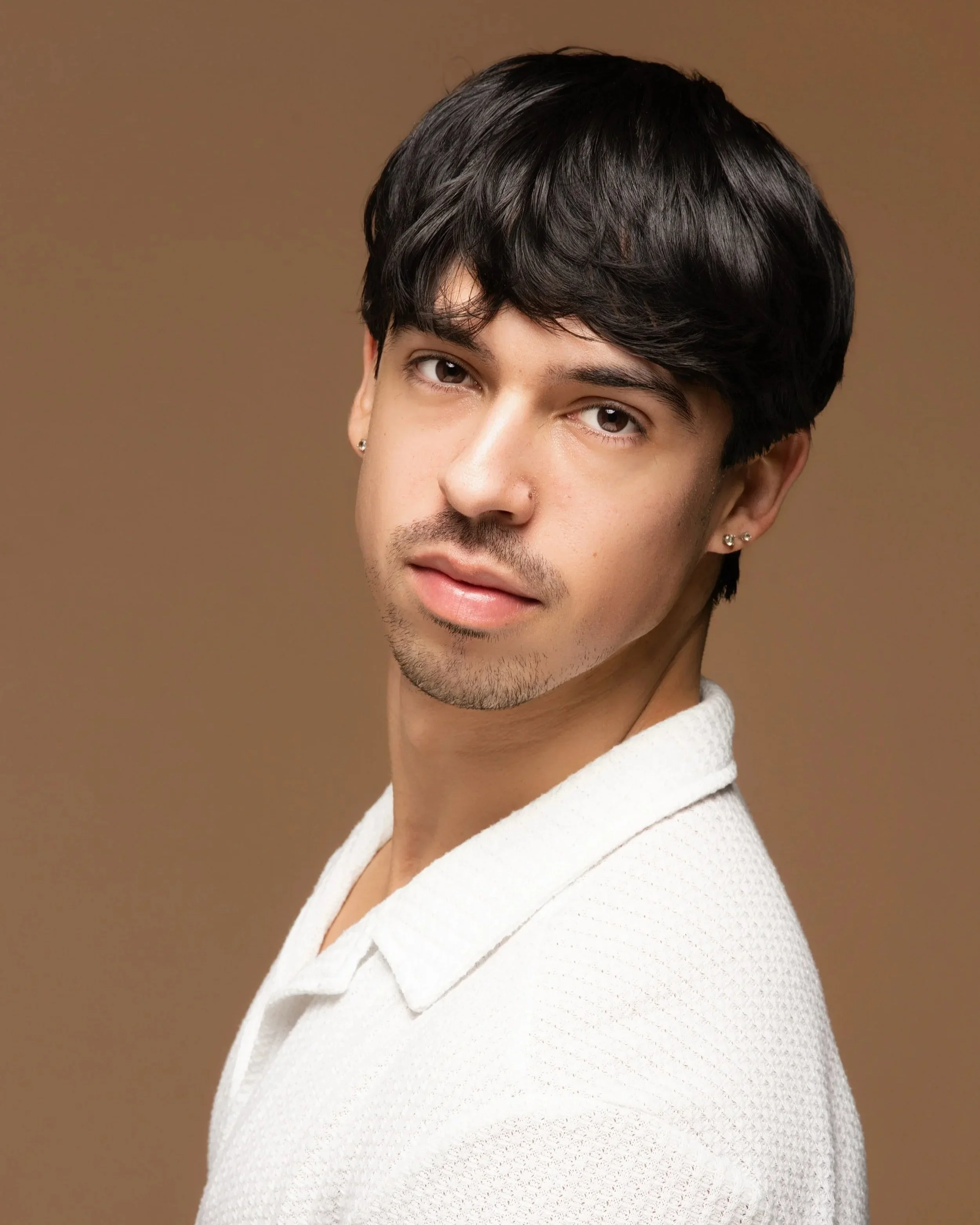Close-up of a young man with dark hair, brown eyes, and earrings, wearing a white textured shirt, standing against a brown background.