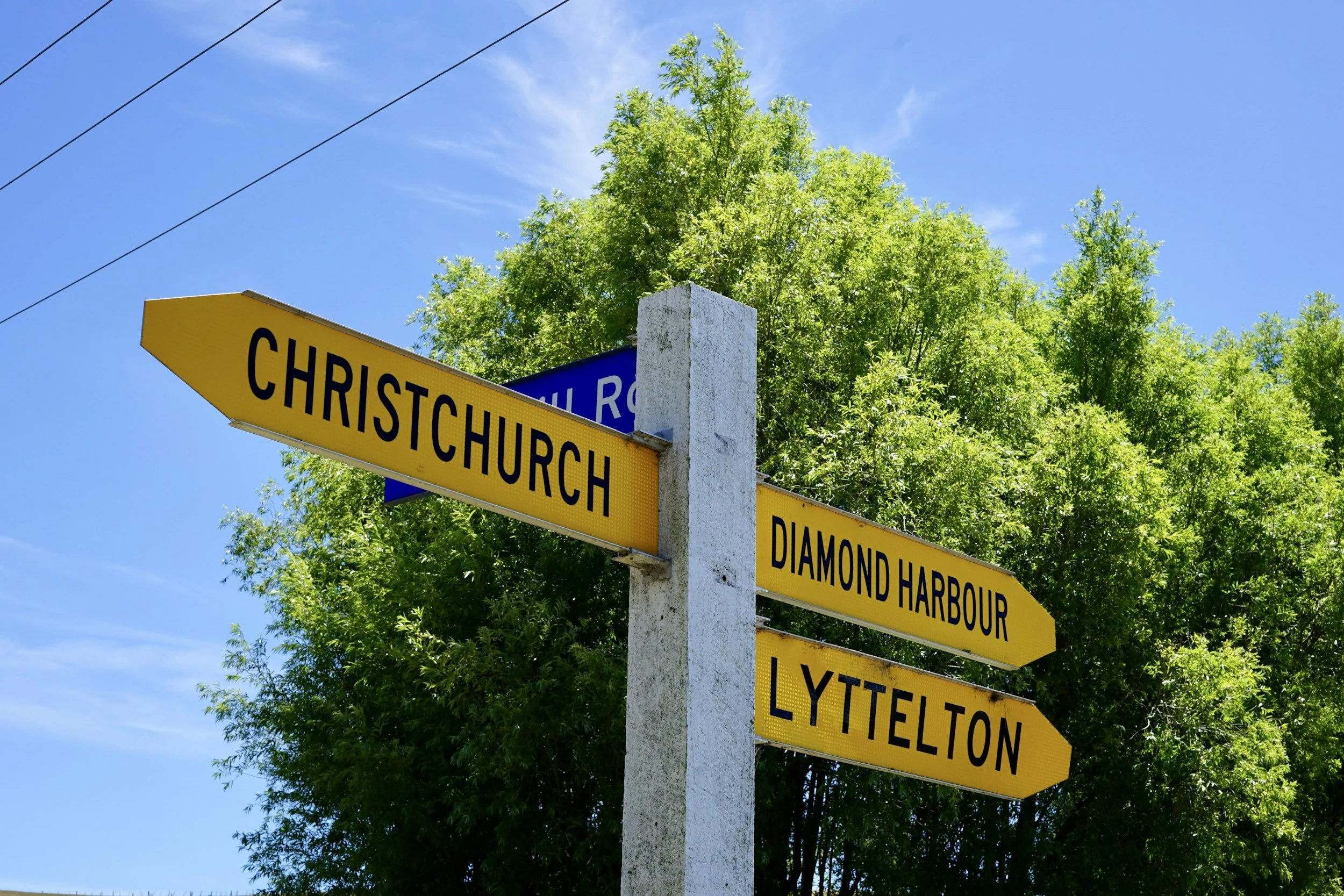 Street signs on a pole, indicating directions to Christ Church, Diamond Harbour, and Lyttelton, with a background of green trees and a blue sky.