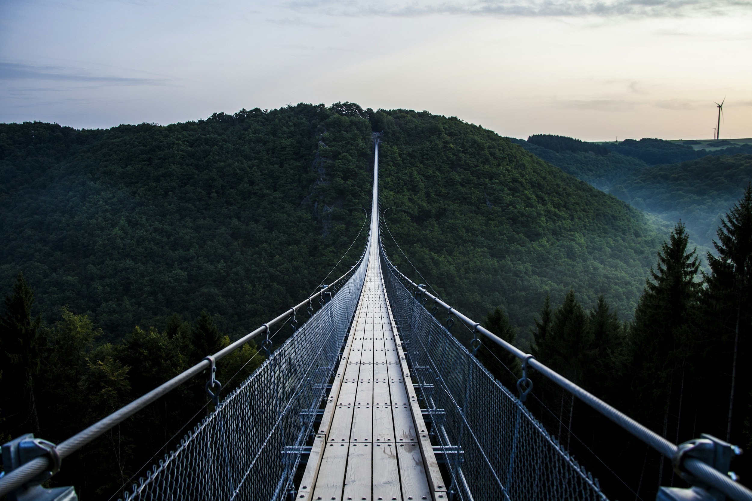 Suspension bridge stretching over a lush green valley with a waterfall on the hillside and wind turbines in the distance.
