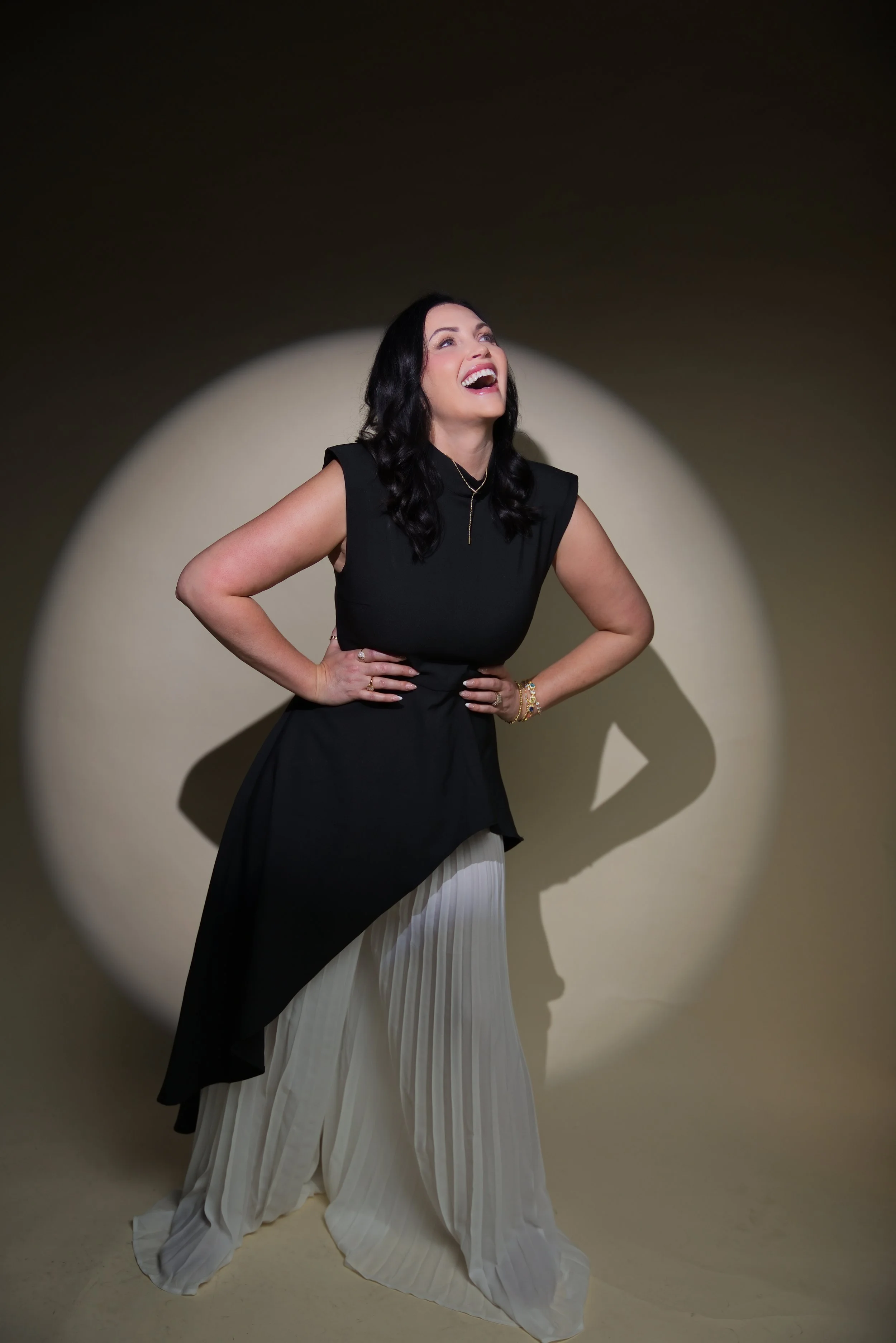 A woman with dark hair styled in loose waves is smiling and looking upward. She is wearing a sleeveless black top, a layered gold necklace, and bracelets. Her hands are on her hips. She is posing in front of a beige background with a spotlight casting a shadow on the wall.