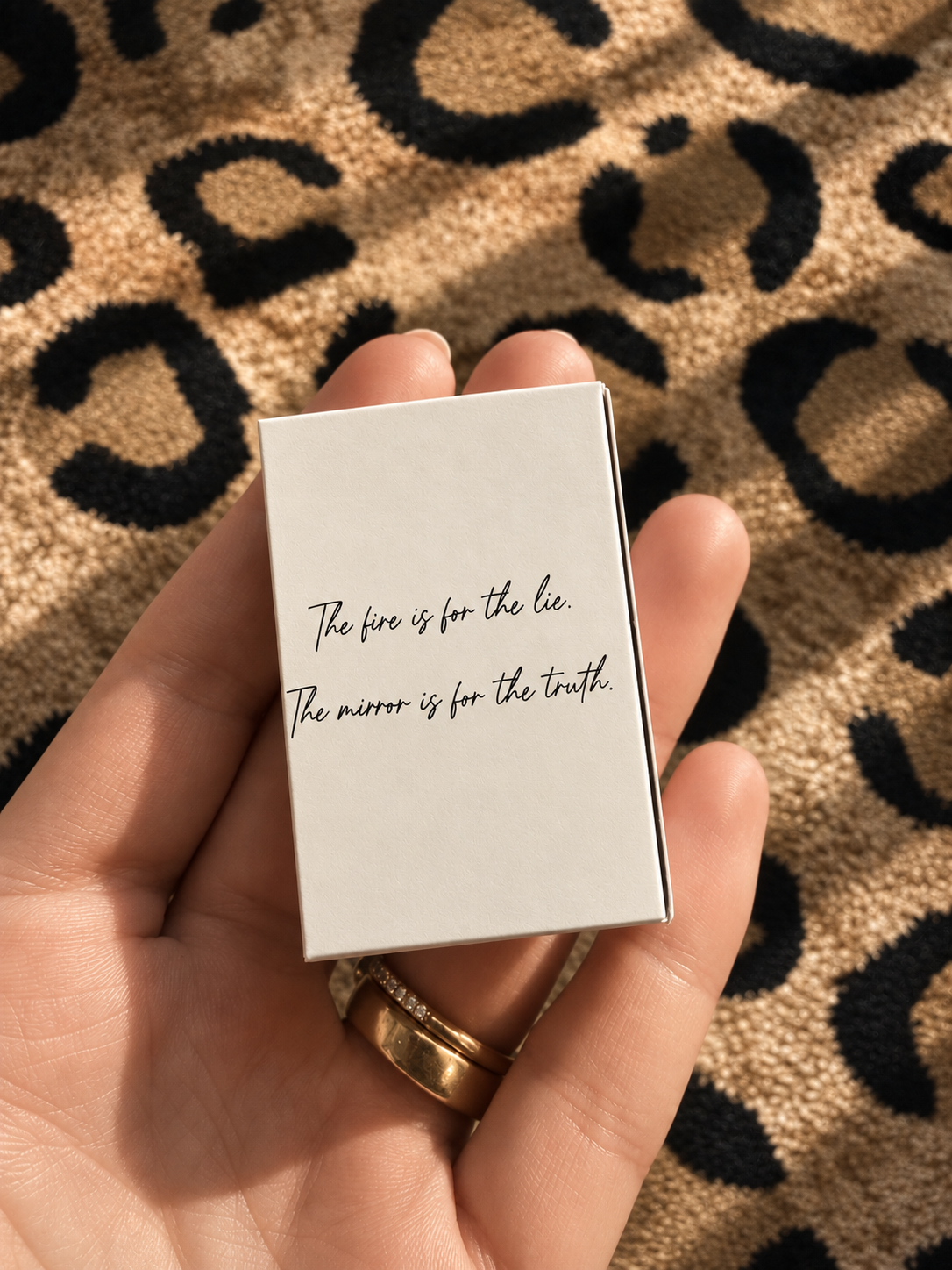 A hand holding a small white box with the quote 'The fire is for the lie. The mirror is for the truth.' written in black cursive text, with a leopard print fabric background.