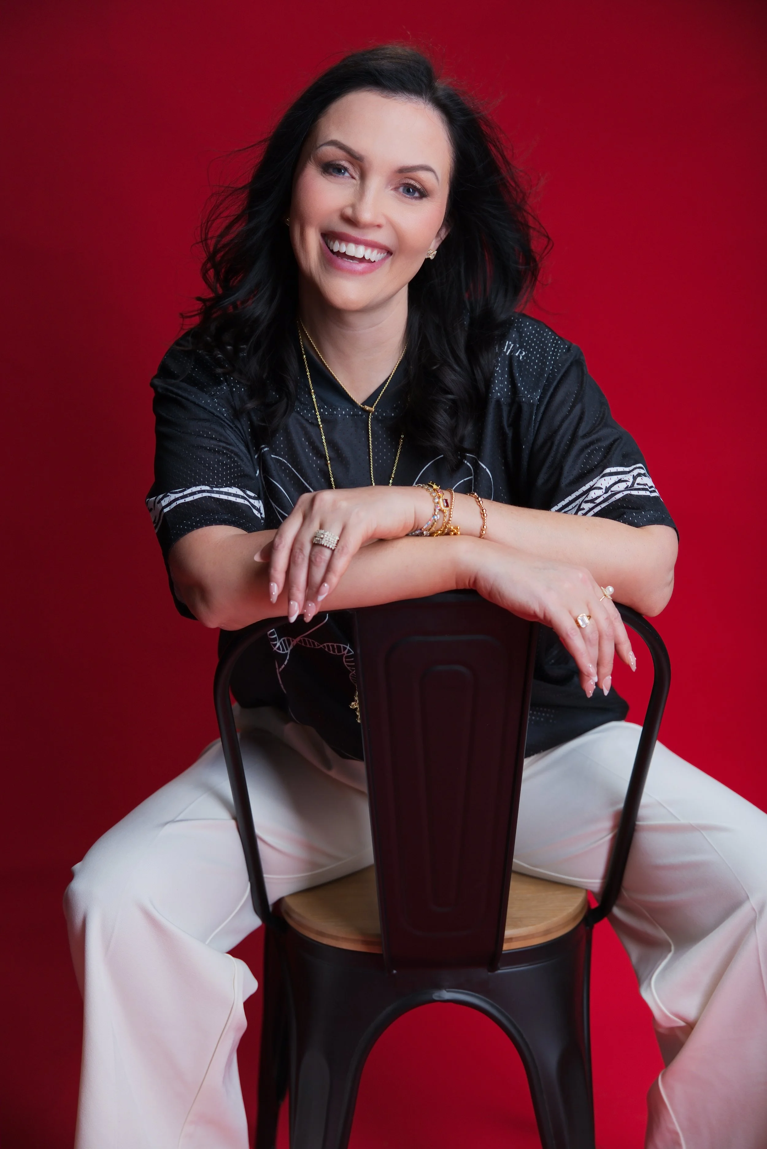 A woman with black hair, wearing jewelry and a black top, is sitting on the floor, leaning on a chair with her arms crossed on the chair's backrest, smiling at the camera against a red background.