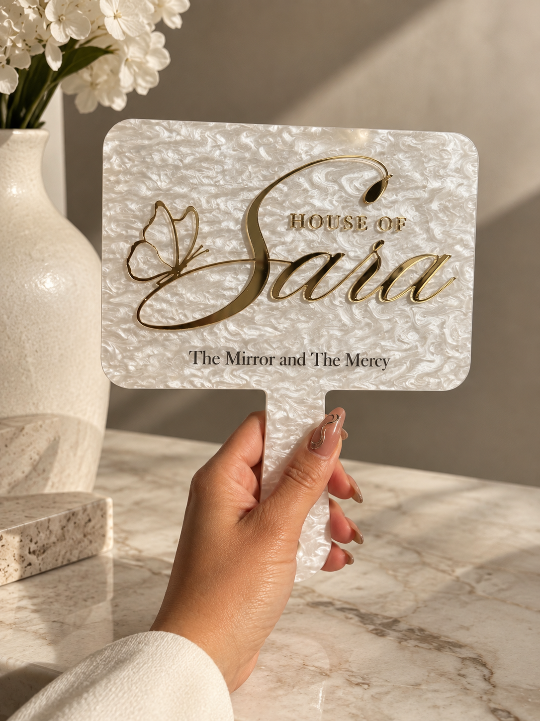A hand holding a sign that reads 'House of Sara, The Mirror and The Mercy' with a butterfly design, placed on a marble table with a beige vase of white flowers nearby.