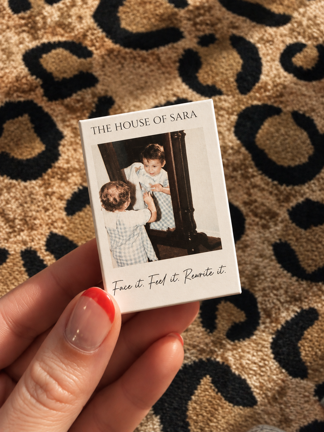 A person holding a small box with a vintage photograph of two children looking at each other in front of a mirror. The box has the title 'The House of Sara' and the tagline 'Face it. Feel it. Rewrite it.' written on it. The background is a patterned carpet with black and tan colors.