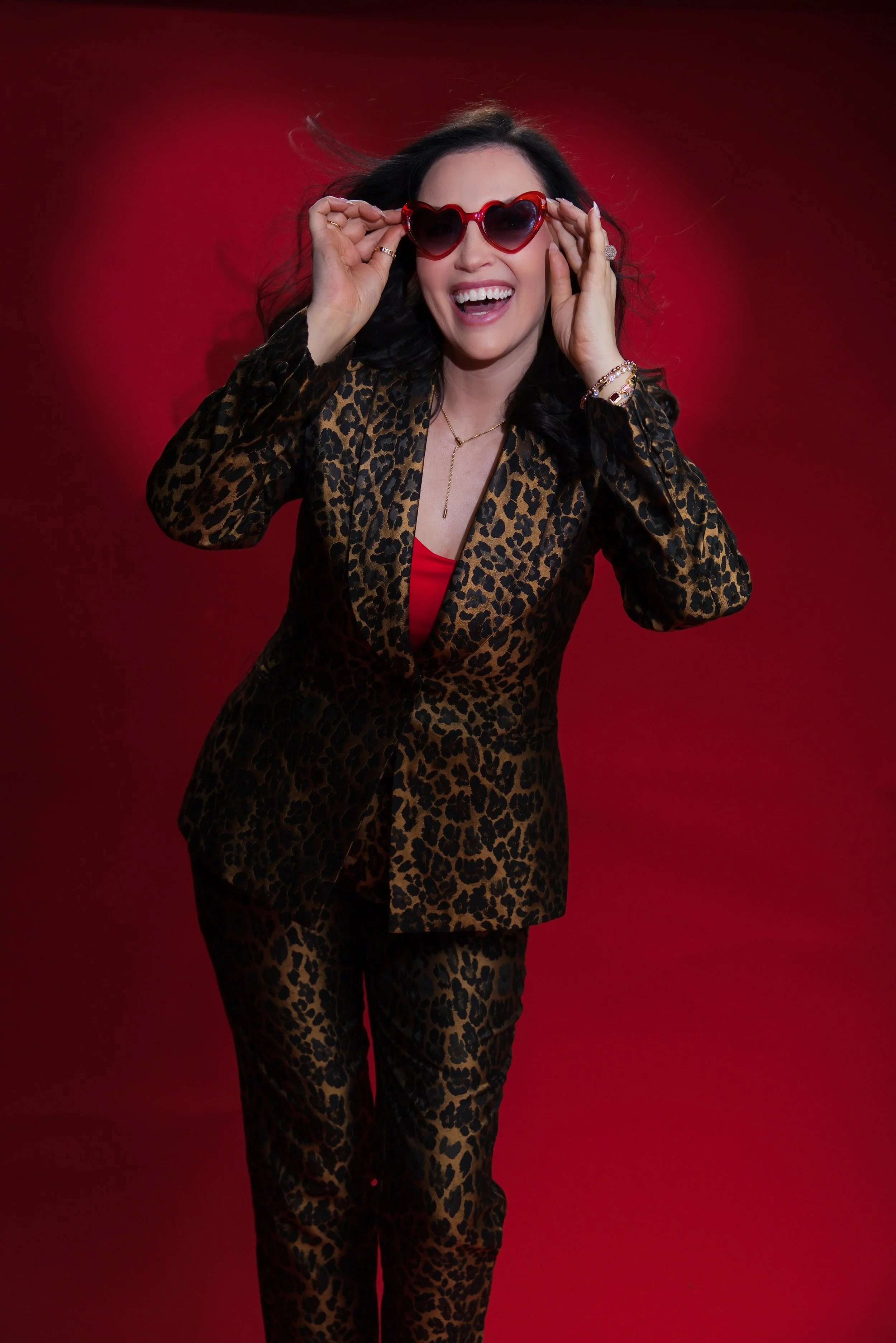 Woman with dark wavy hair wearing leopard print suit, red top, and heart-shaped sunglasses, smiling against a red background.