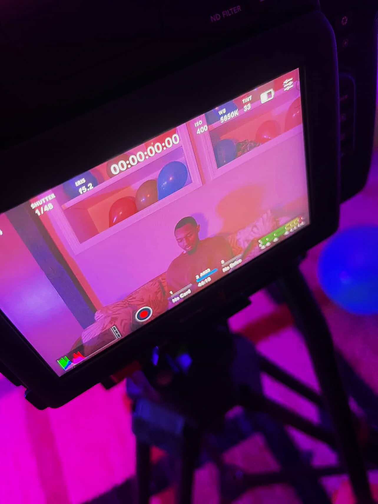 A camera monitor displaying a video recording of a man sitting on a bed in a room with pink walls and balloons on a shelf.