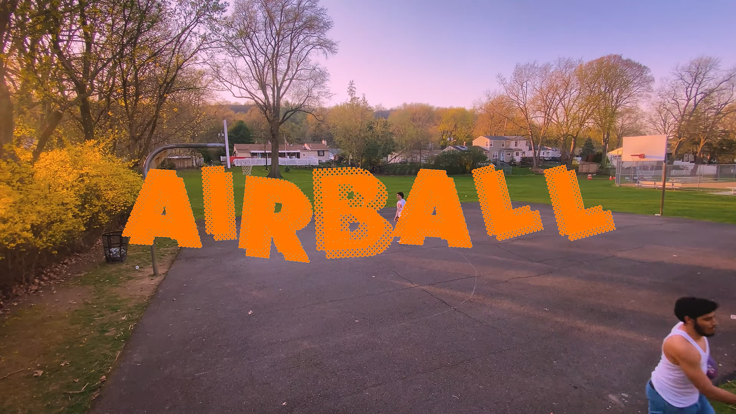 A park scene at sunset with a basketball hoop on the left and a large orange sign spelling 'ARUBA' in the foreground. There are trees with autumn foliage, houses in the background, and a person in a white tank top walking at the bottom right.