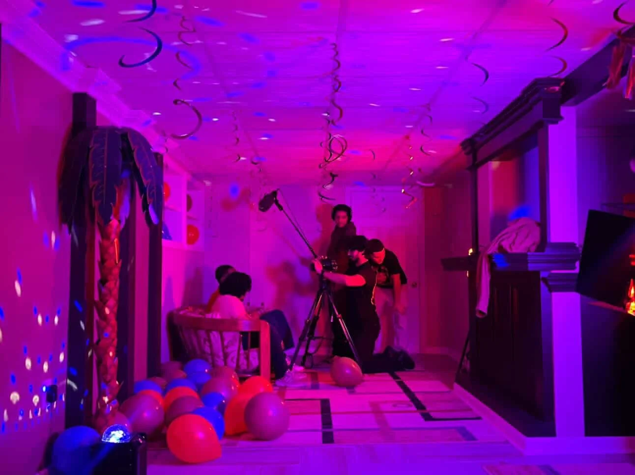 Indoor party scene with purple and pink lighting, streamers hanging from the ceiling, several balloons on the floor, a woman sitting on a chair, two men adjusting camera equipment, and a small group of people in the background.