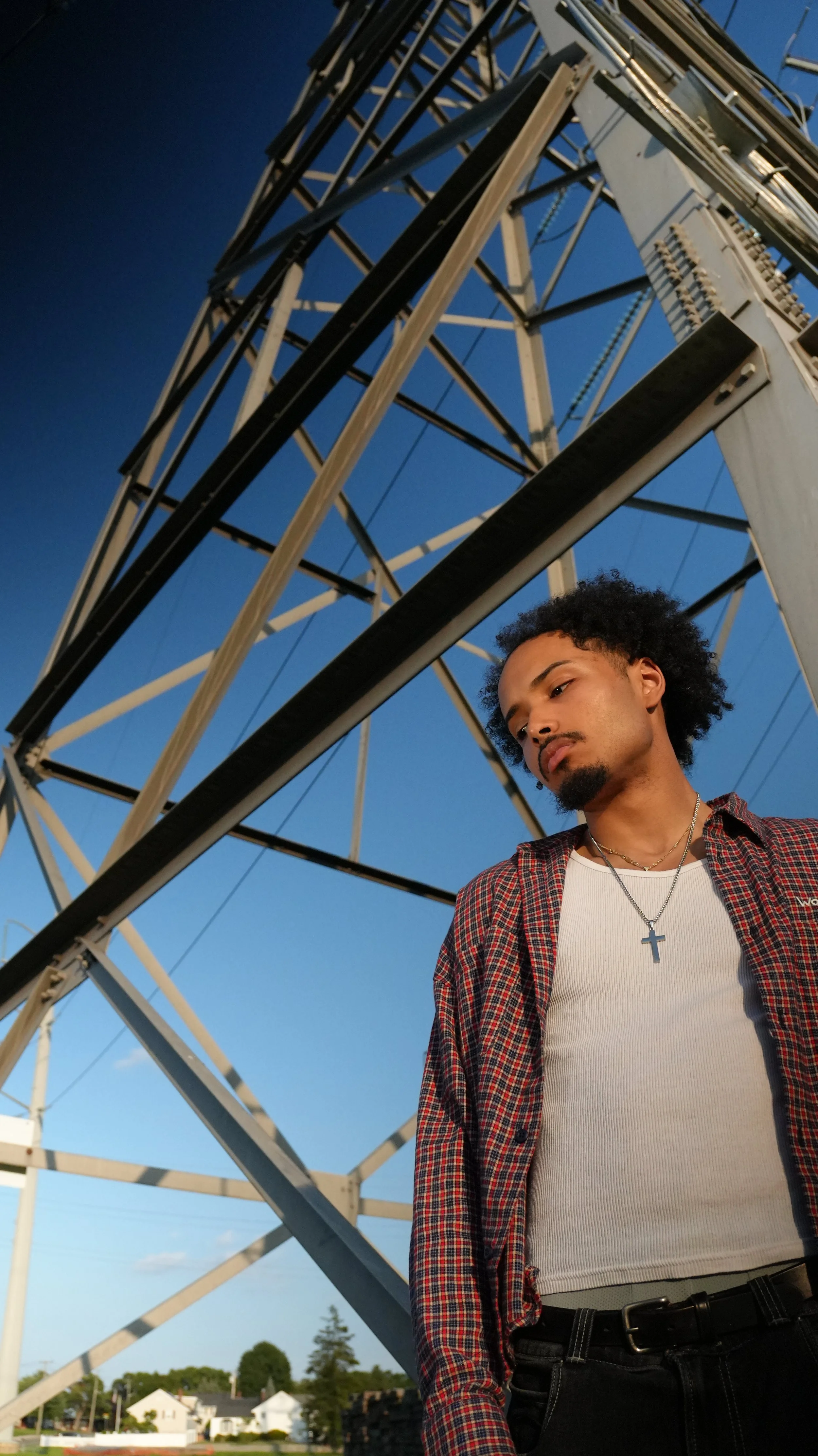 A young man with curly hair and a goatee wearing a cross necklace, a white t-shirt, a red plaid shirt, and black jeans standing near a large electrical tower under a clear sky.