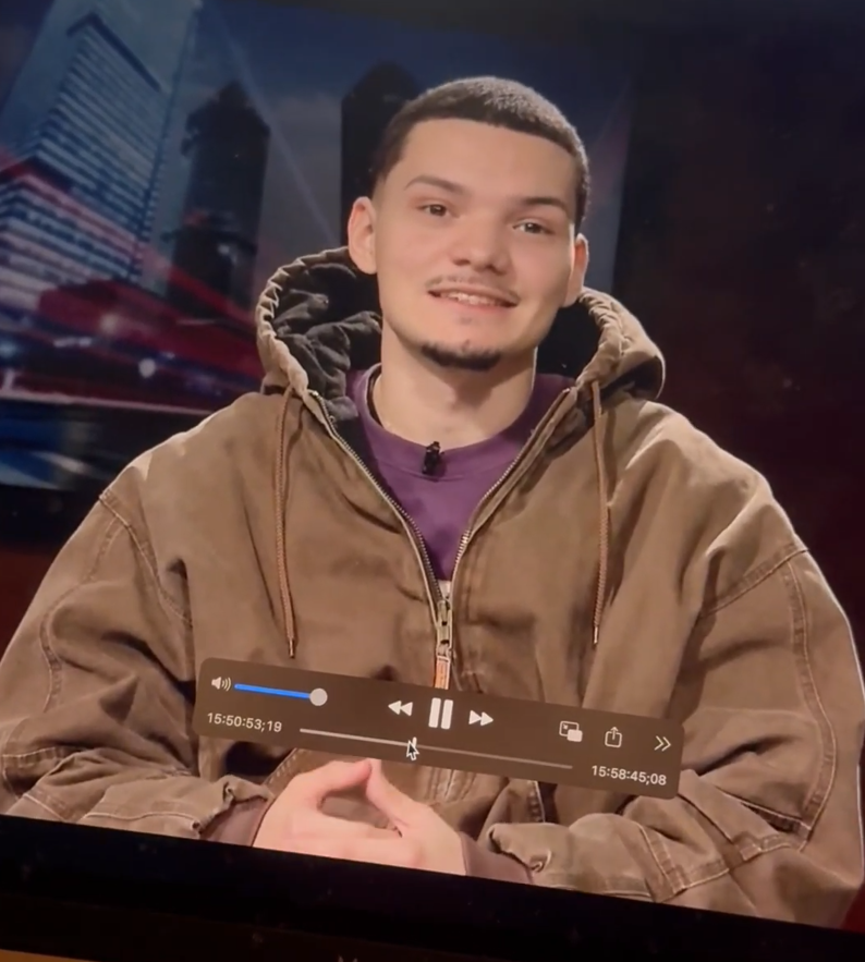 Young man with short dark hair, goatee, wearing a brown hoodie over a purple shirt, sitting at a table in front of a cityscape background.