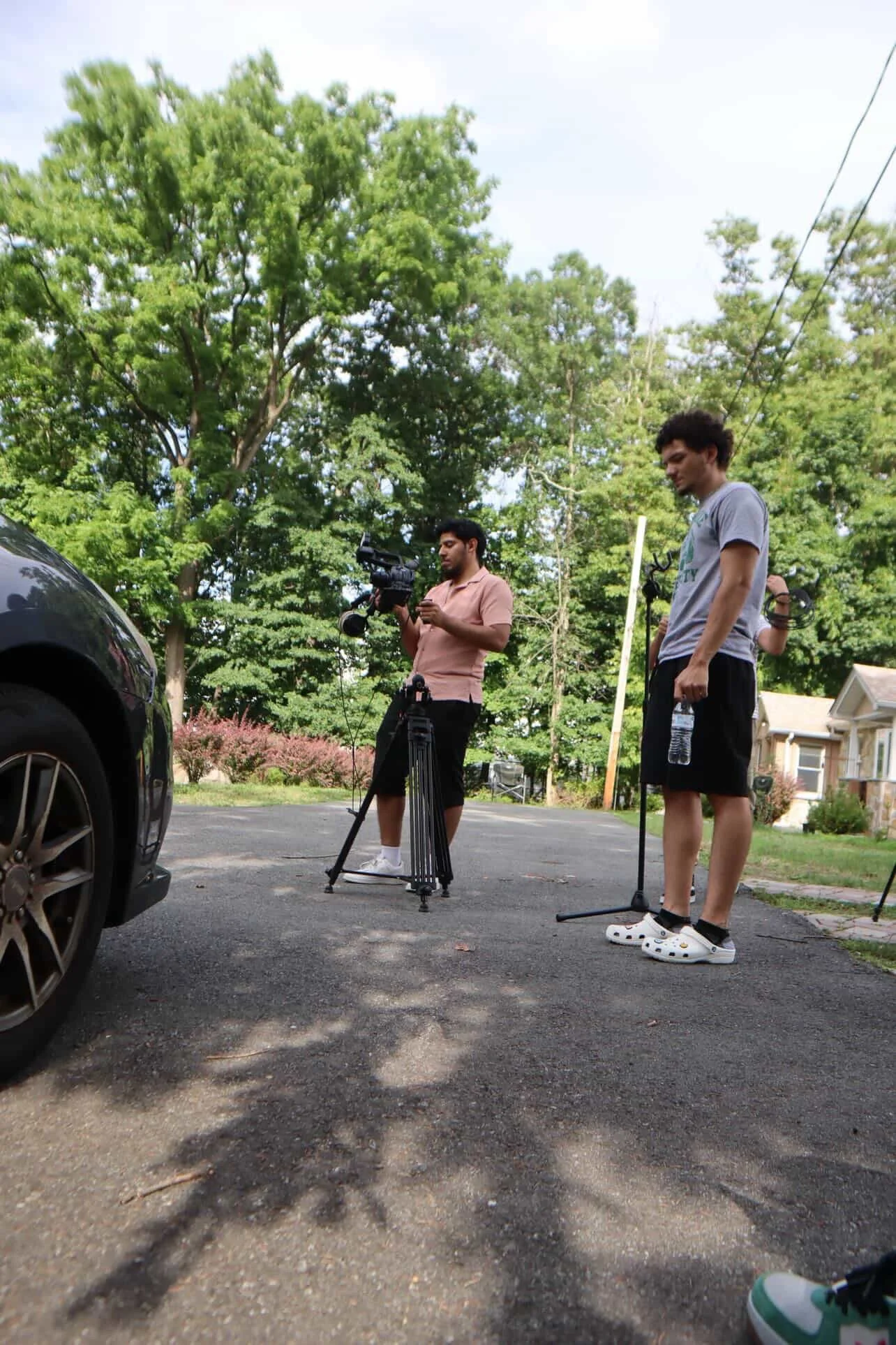 Two men stand on a driveway outdoors, one operating a video camera on a tripod and the other holding a water bottle, with trees and houses in the background.