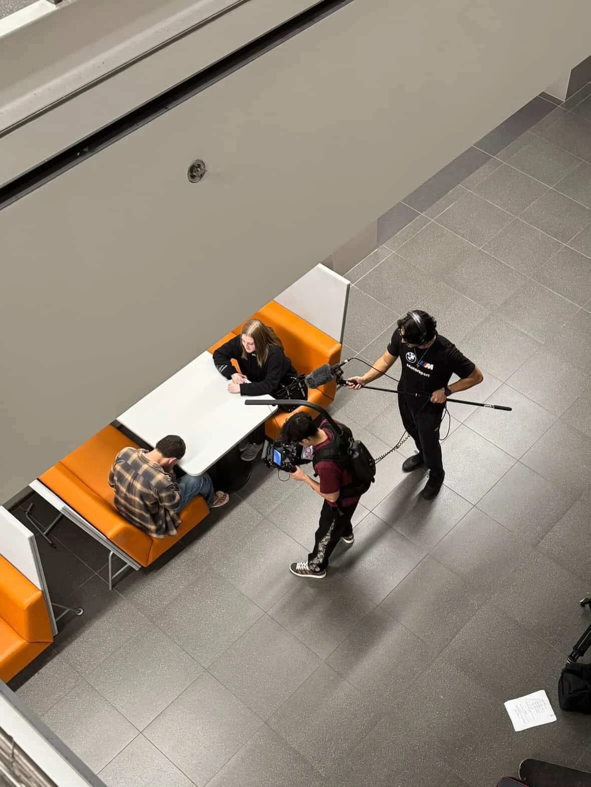 A film crew making an interview at a table in a modern, indoor public space with gray tile flooring. One person sits at the table, another on the booth, and a third person operating a camera, while a crew member holds a boom microphone.