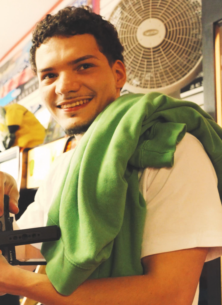 A young man with curly dark hair smiling while holding a Sony camera. He has a green hoodie draped over his shoulders and wears a light-colored T-shirt. A fan is visible on the wall behind him.