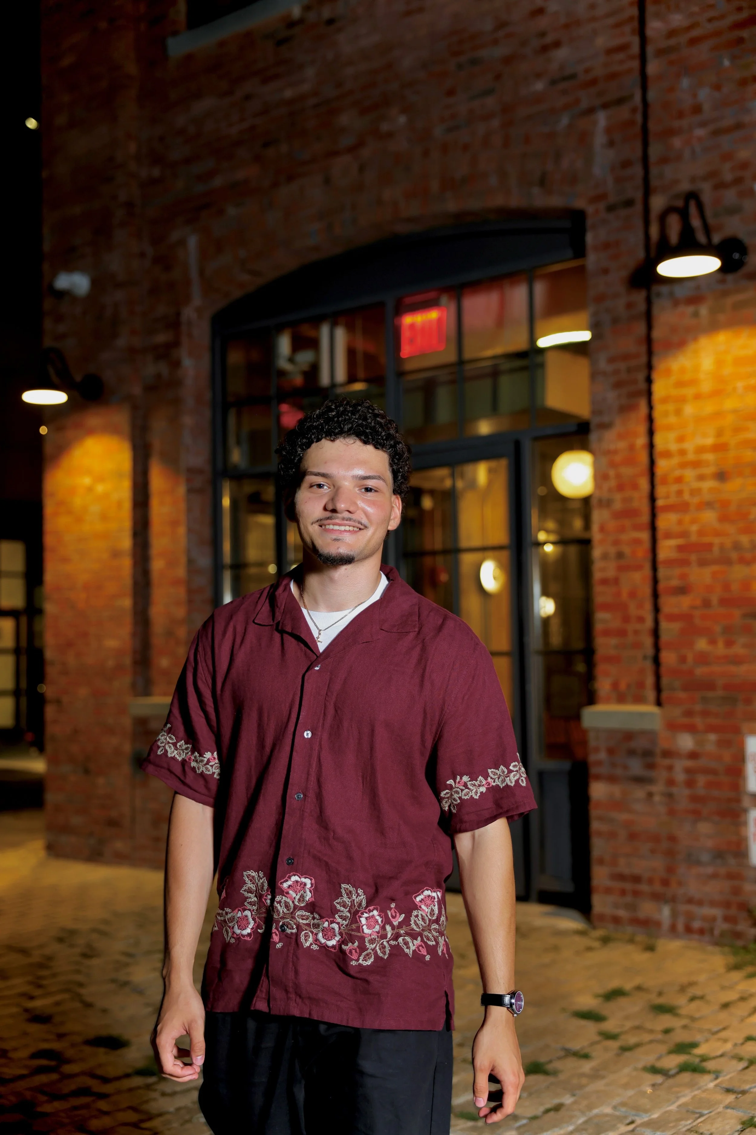 A young man with curly dark hair and light skin smiling, standing outside in front of a brick building with large windows and warm lighting, wearing a burgundy embroidered shirt, black pants, and a watch on his left wrist.