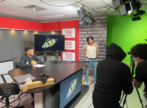 Two people conducting a video interview or recording in a studio with a green screen and studio lights, while a person sits at a desk with a laptop and a camera on the table.