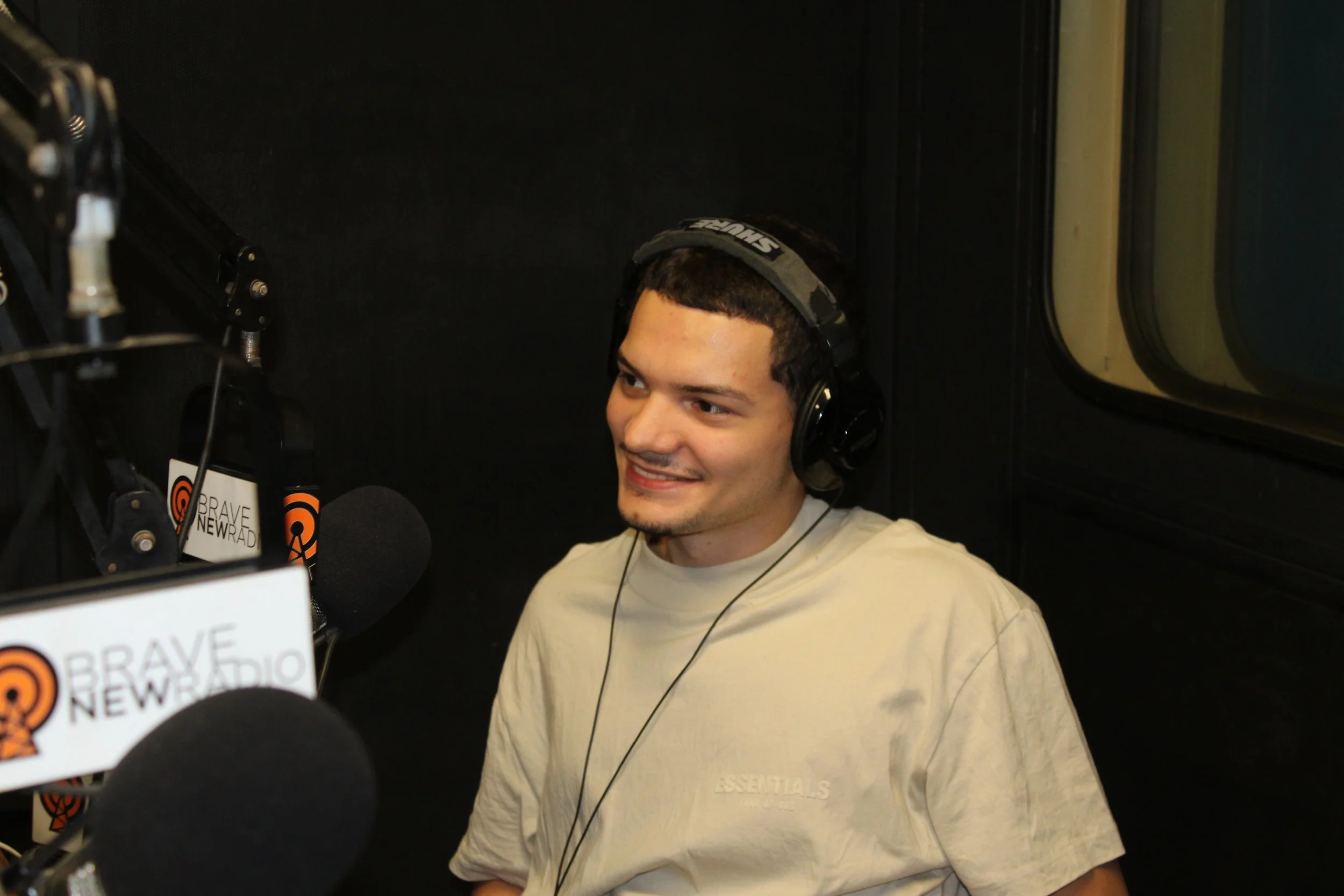 Young man wearing headphones, sitting in a recording studio with multiple microphones and soundproofing, smiling during a radio interview.