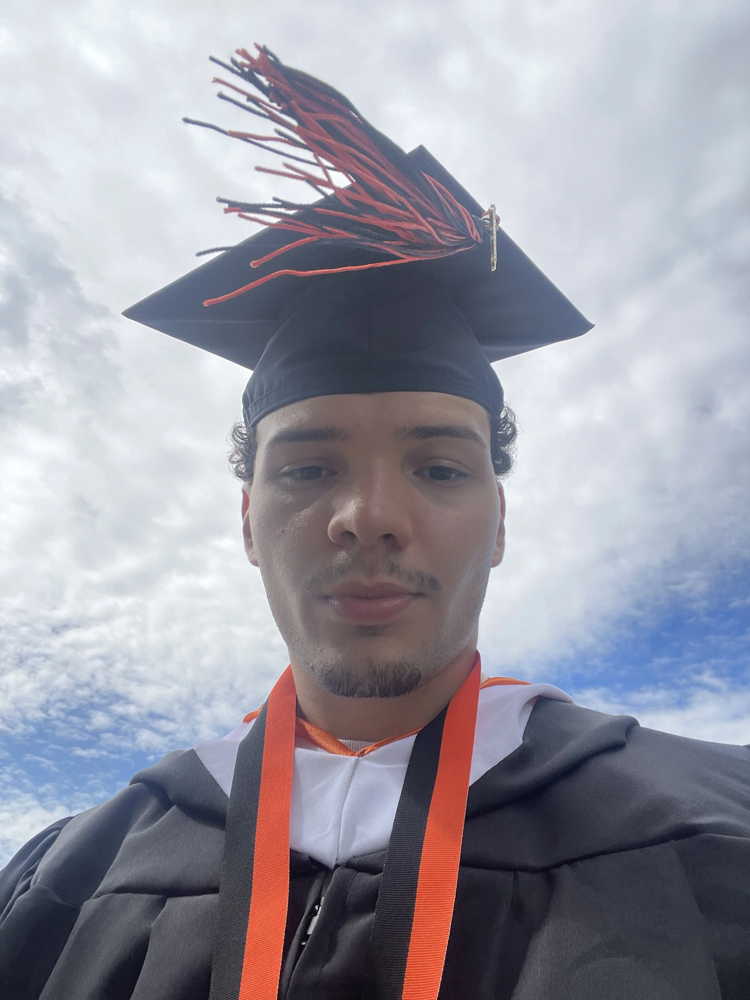 A young man wearing a graduation cap, gown, and orange medal, outdoors against a cloudy sky.