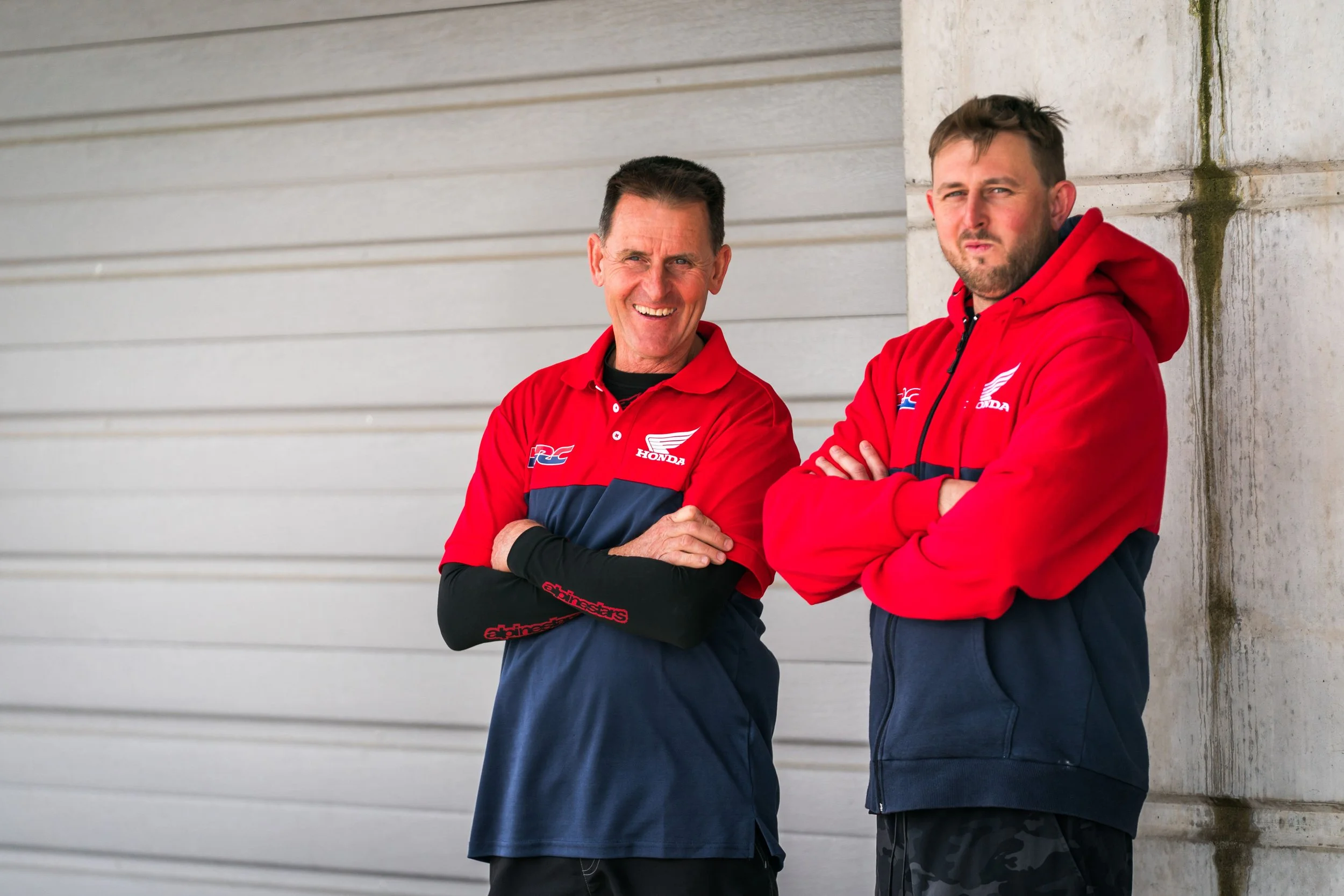 Two men in red Honda racing jackets standing against a grey wall with metal siding. They are smiling and look confident.