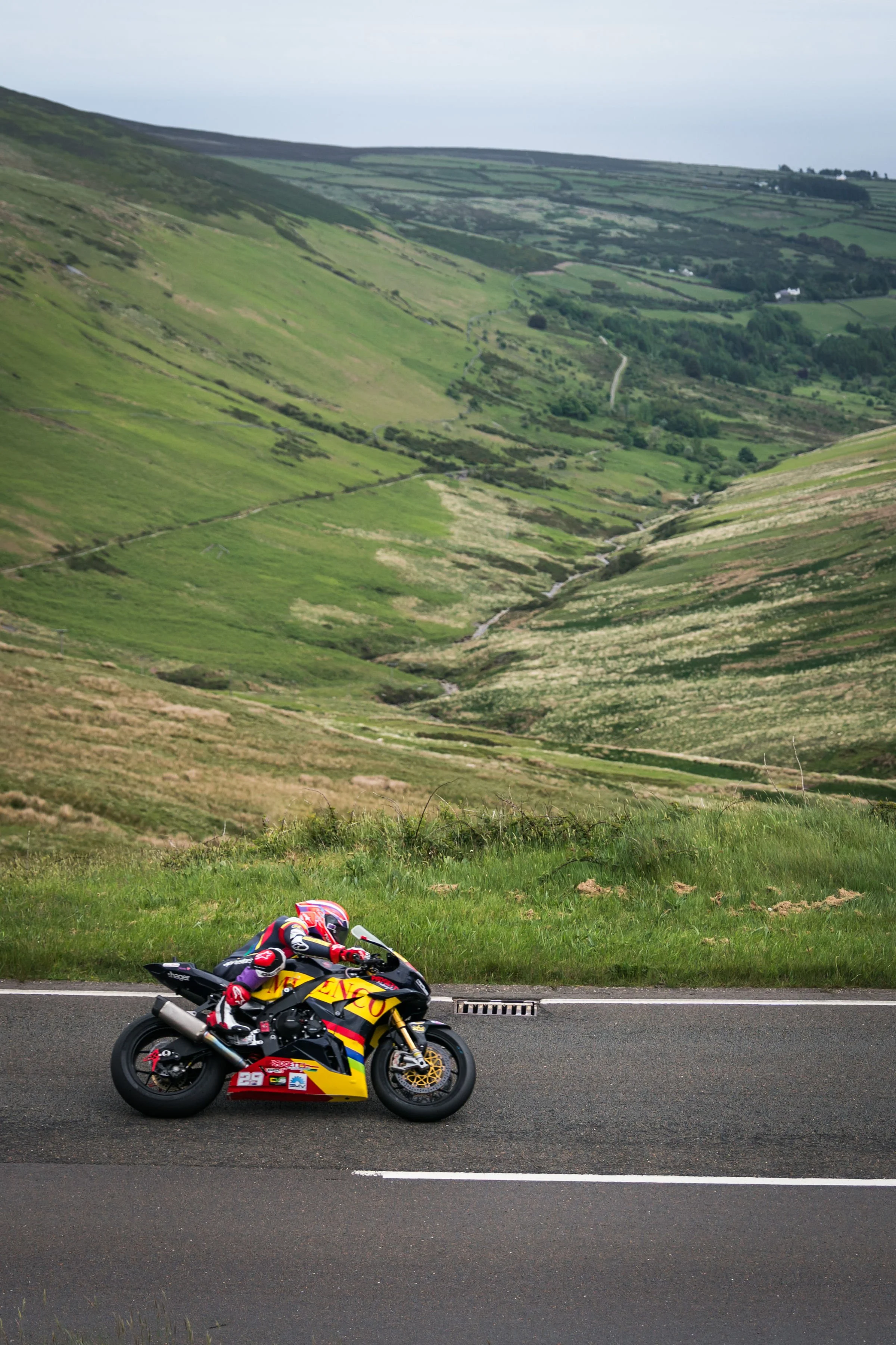 A motocross racer wearing a colorful helmet and racing suit riding a yellow and red motorcycle on a paved road with green hills in the background.