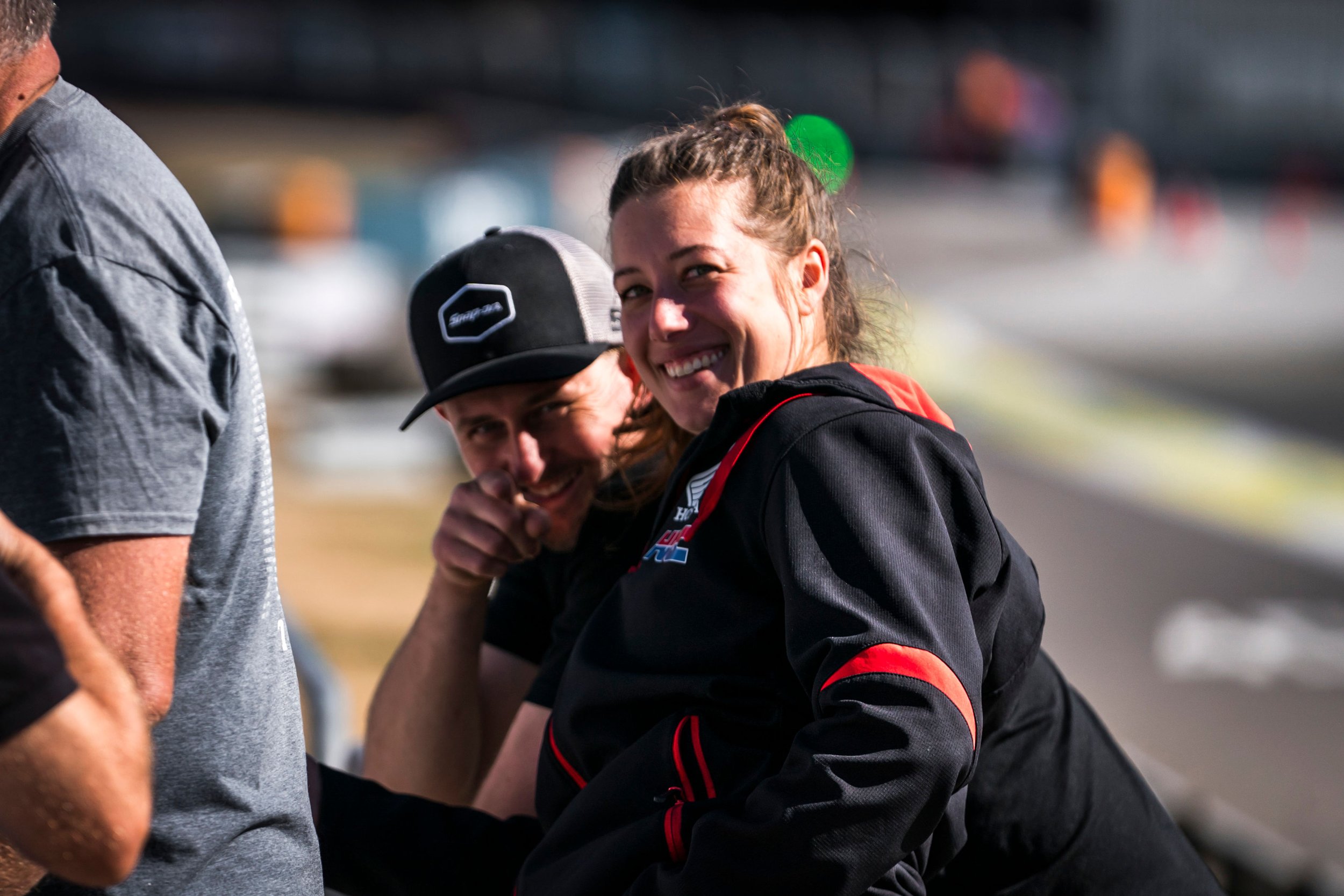 Smiling woman in racing jacket flexing her arm at a sporting event, with a man wearing a cap standing nearby, face partially visible.