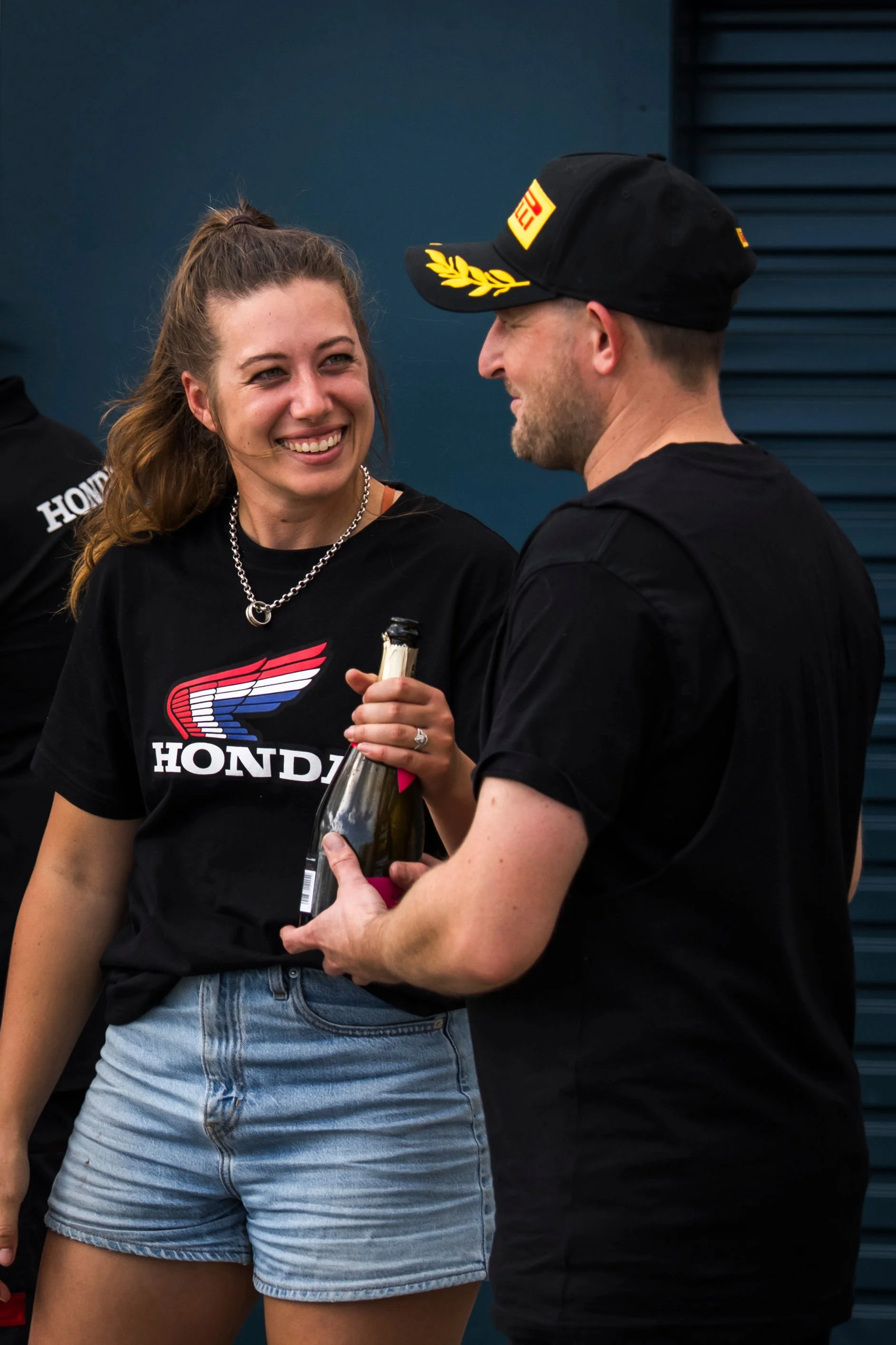 A woman with brown hair in a ponytail, wearing a black Honda T-shirt and denim shorts, is smiling while holding a bottle. She is talking to a man wearing a black cap with yellow details and a black shirt.