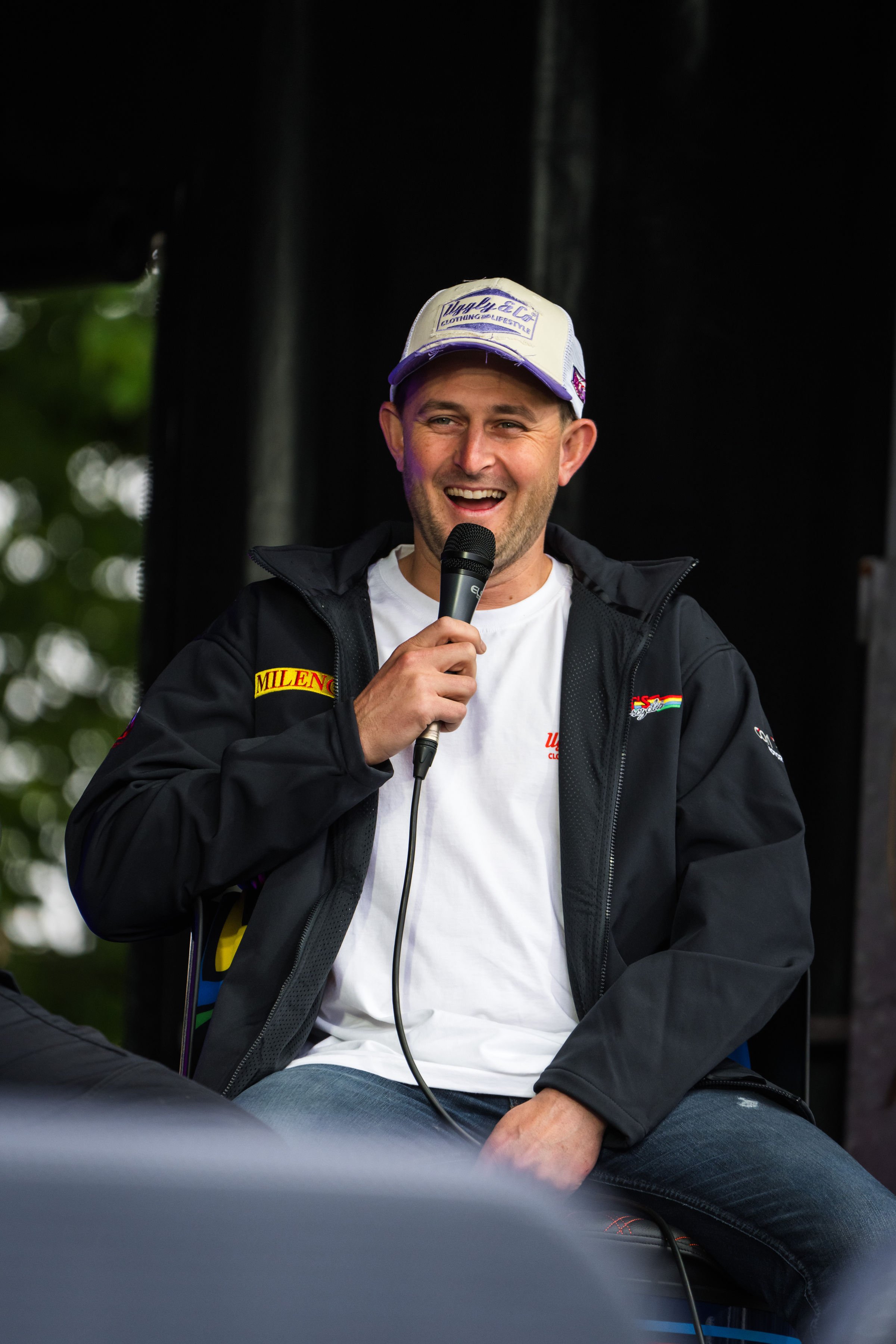A man holding a microphone, smiling, wearing a white cap with purple accents, and a black jacket with various patches, sitting outdoors with blurred greenery in the background.