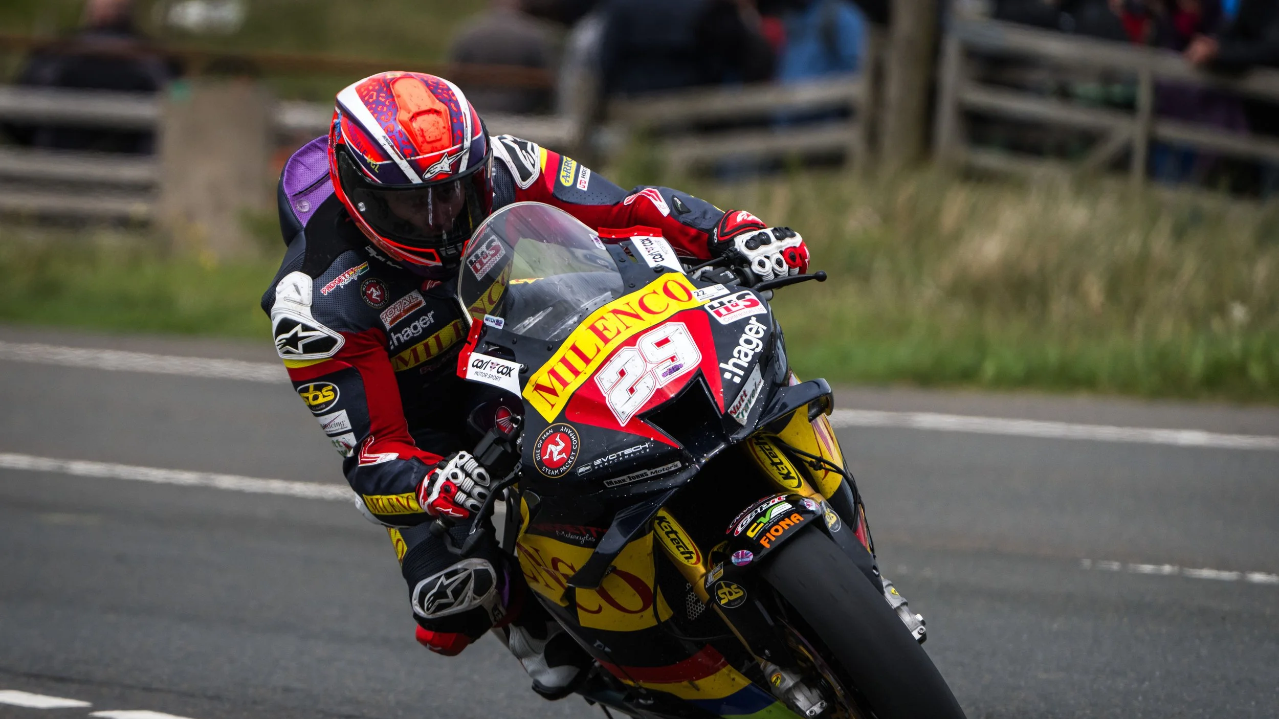 Motorcycle racer leaning into a turn on a race track, wearing a colorful helmet and racing gear with sponsor logos.