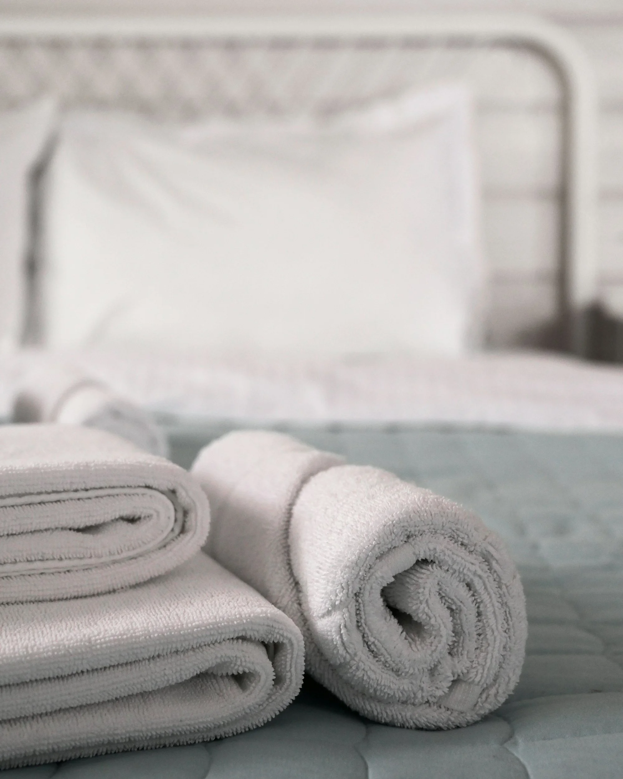 Four folded white towels stacked on top of each other on a bed or table with a gray quilted cover in the background.