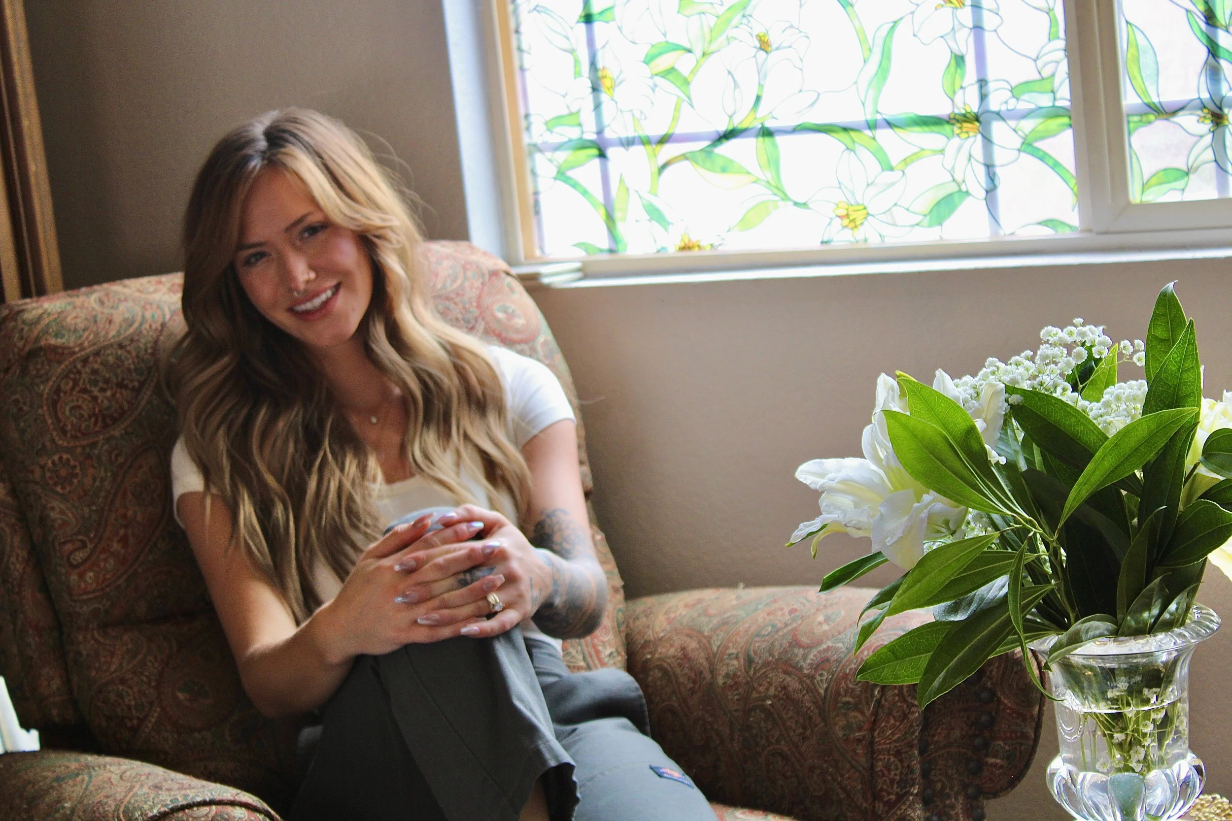 A woman with long blonde hair and tattoos on her arms relaxing on a patterned sofa, holding a mobile phone, sitting near a window with a stained glass floral design, next to a large glass vase filled with white flowers and green leaves.