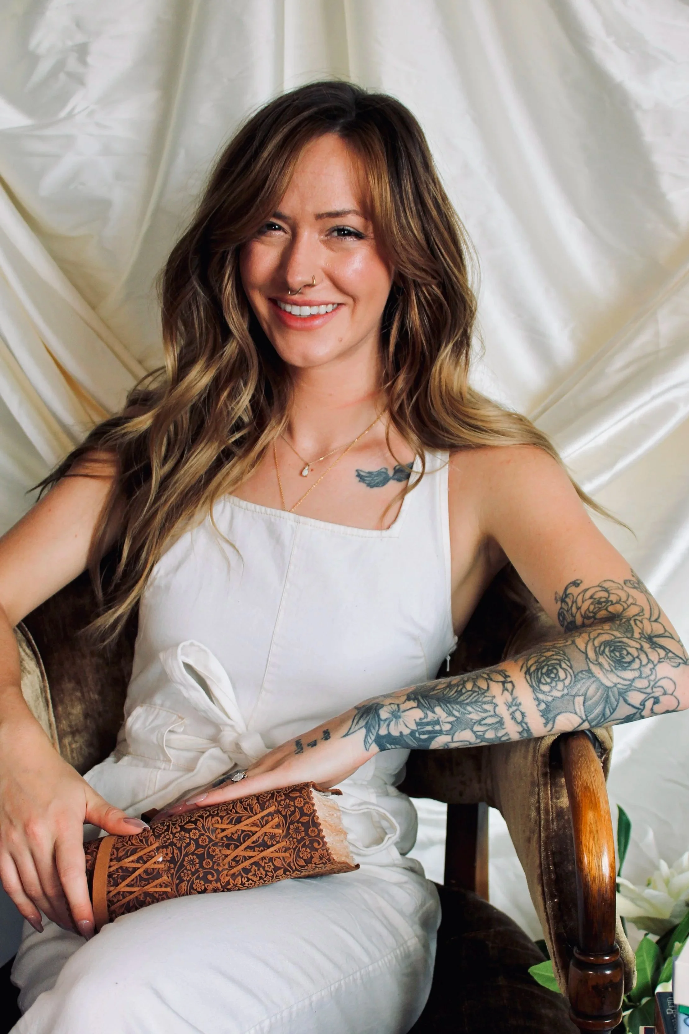 A smiling woman with shoulder-length brown hair and tattoos on her arm, sitting on a vintage wooden chair with a white silk background. She is wearing a white sleeveless top and has a necklace with a small pendant.