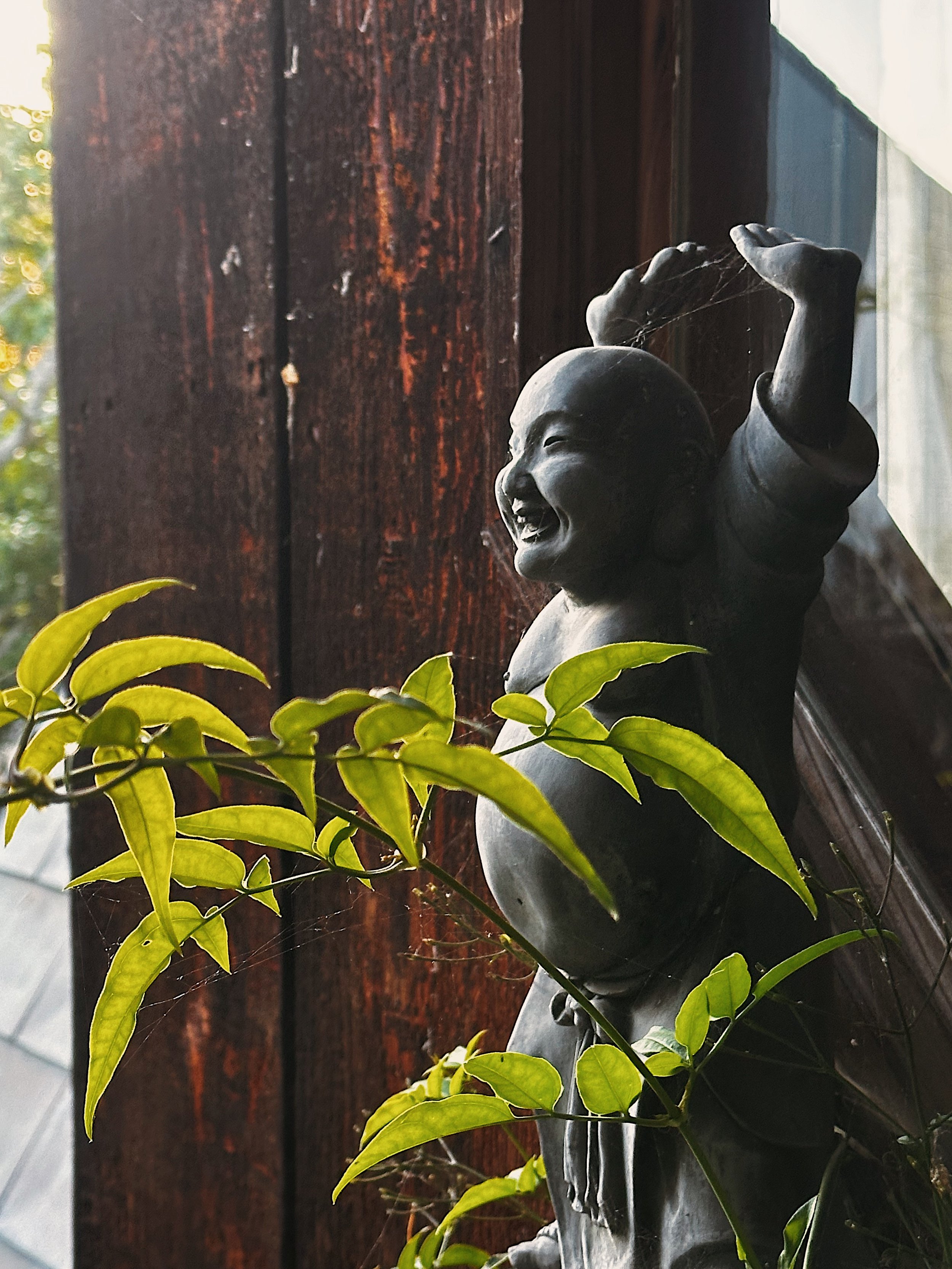 A small statue of a smiling child holding a string, surrounded by green leaves, on a dark wooden surface with cobwebs.